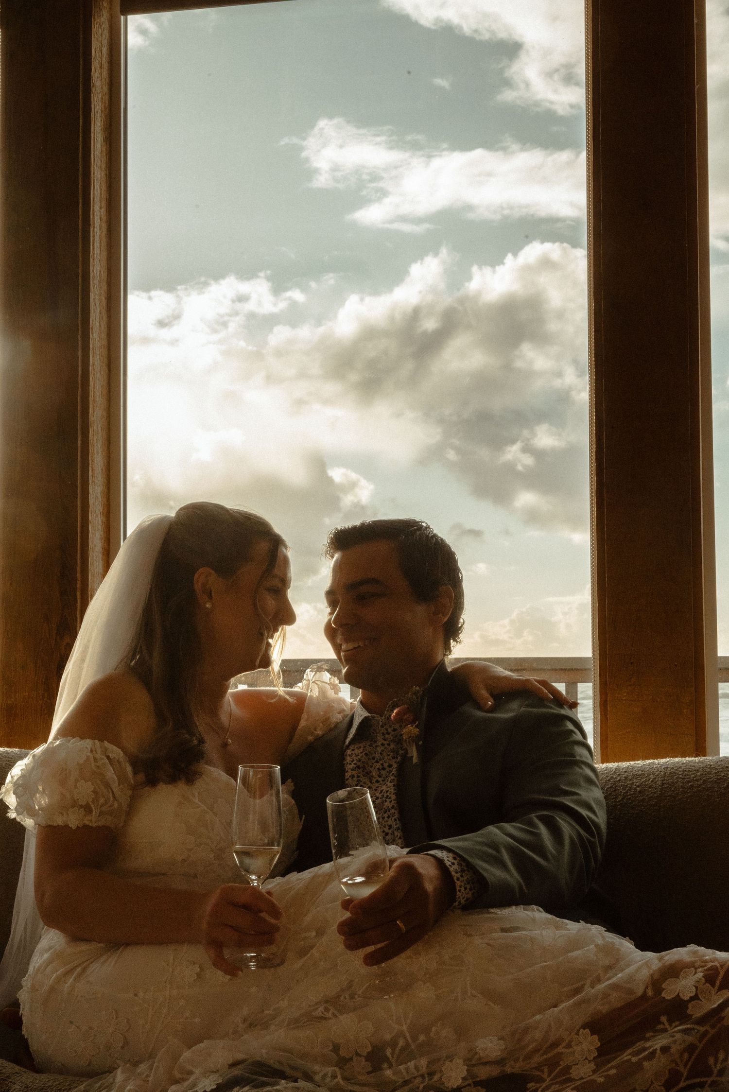Bride and groom sitting together holding champagne glasses, smiling at each other with a cloudy sky visible through large windows.