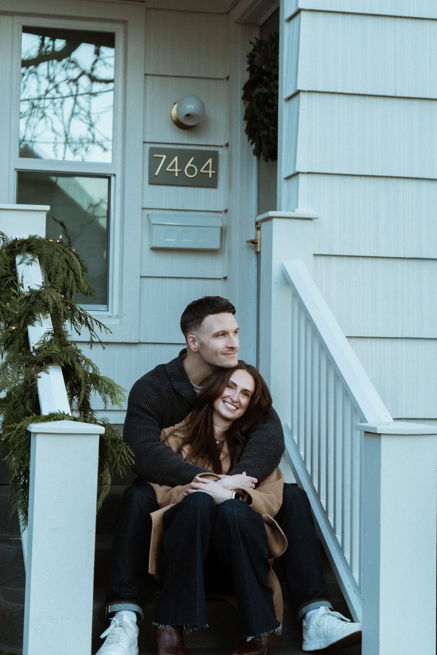 A couple sits on front porch steps of a house numbered 7464, embracing warmly with holiday greenery decorations nearby.