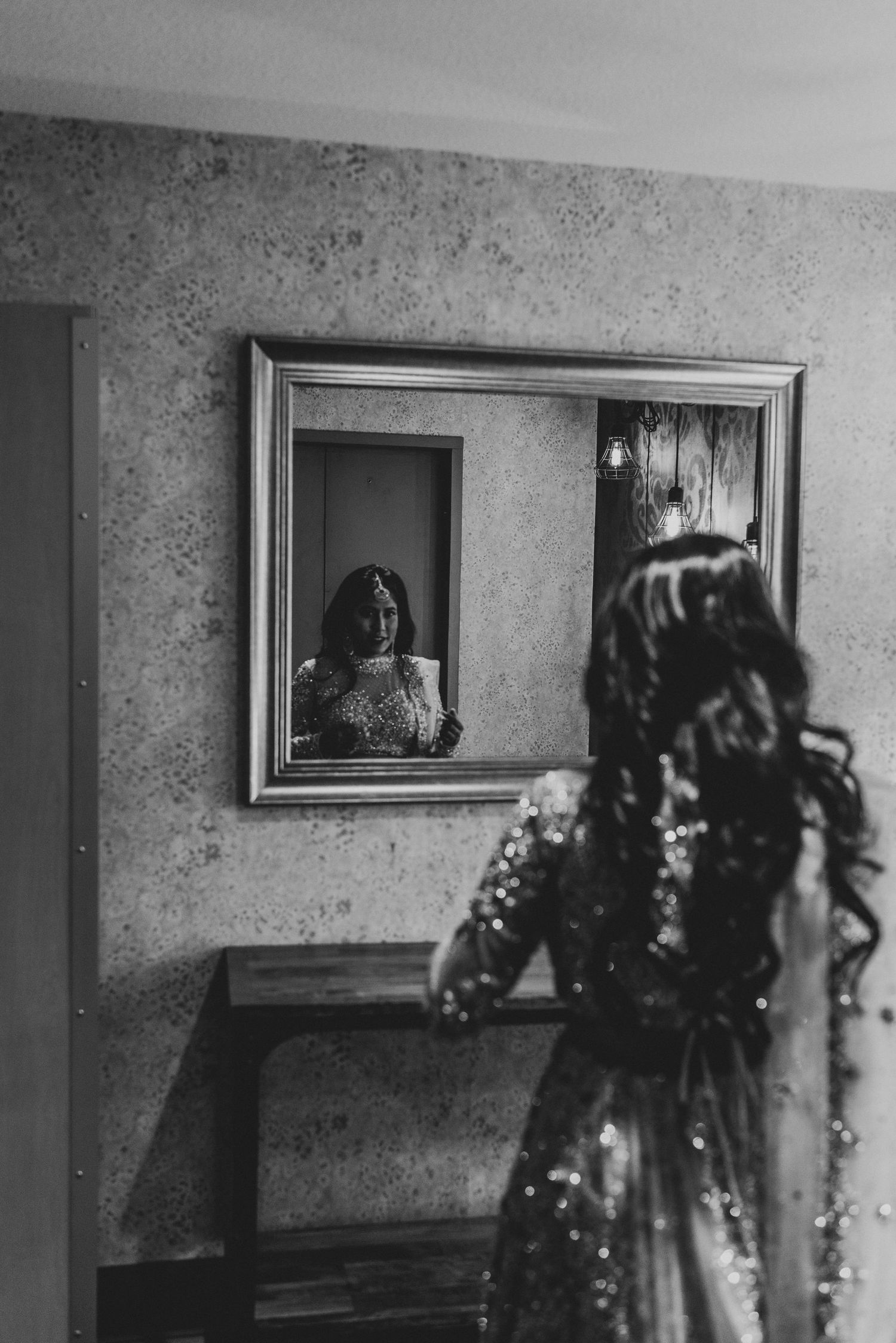 Black and white photo of a bride in sequined dress viewing her reflection in an ornate framed mirror.