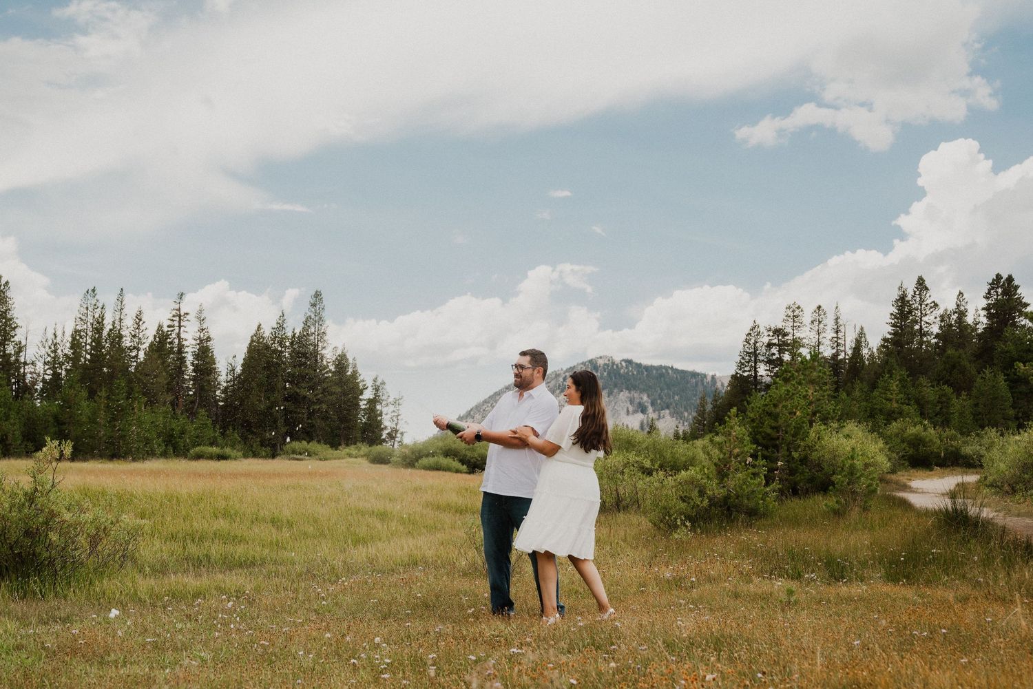 Couple dancing in a wildflower meadow with pine trees and mountains under a cloudy sky.