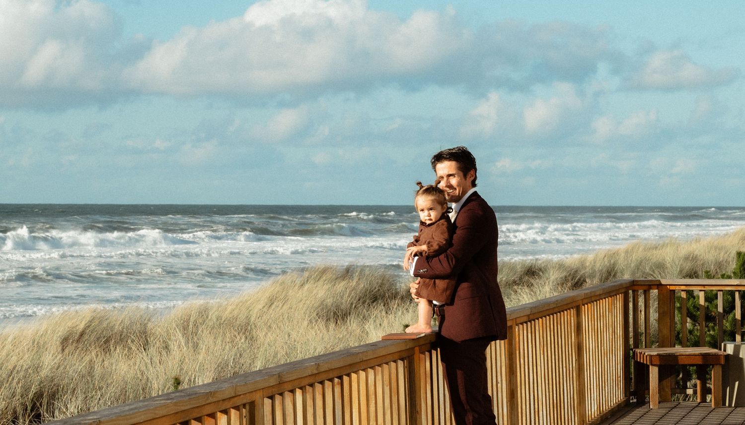 A man in a dark suit holds a young child while standing on a wooden boardwalk overlooking dune grass and ocean waves under a cloudy sky.