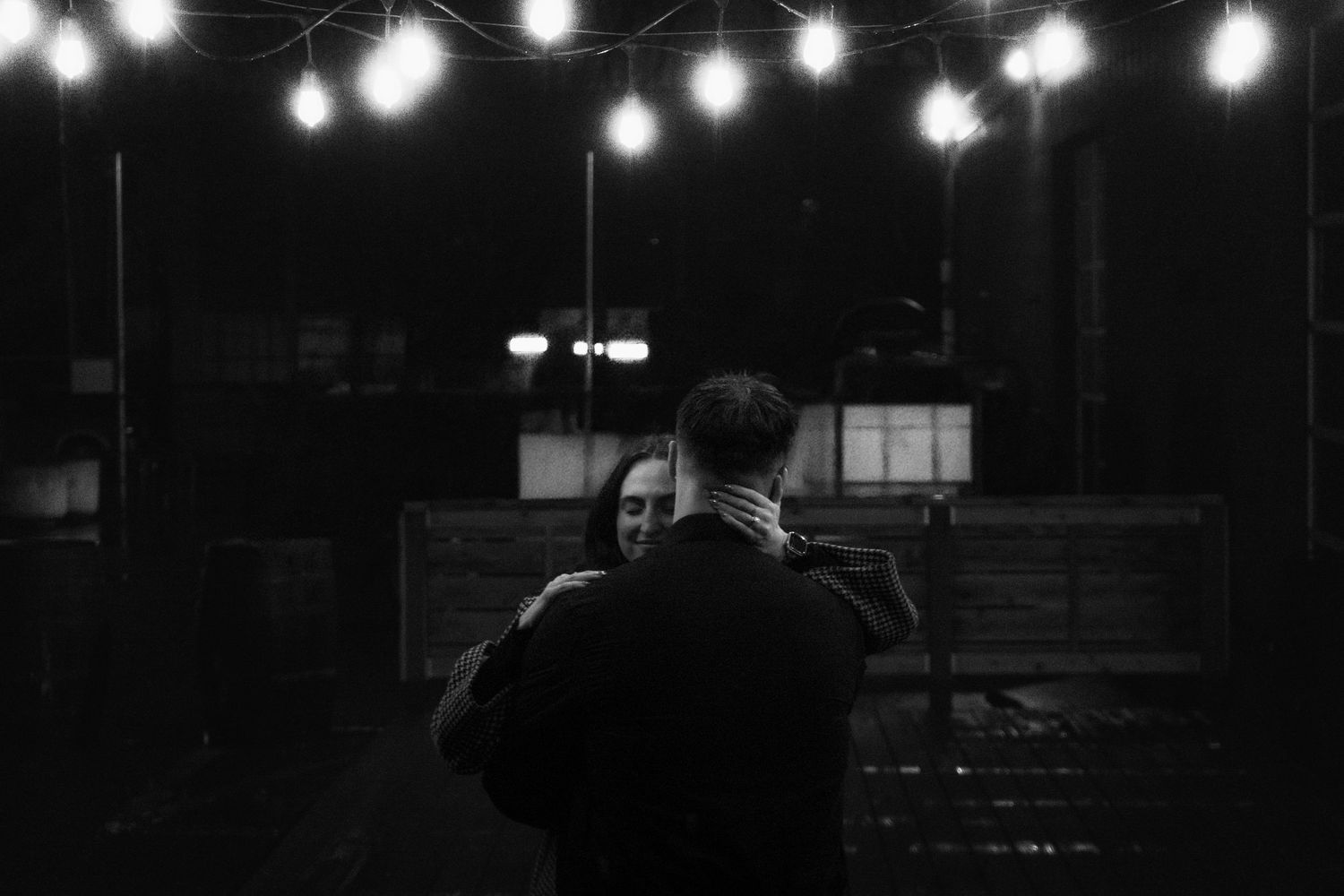 Black and white photo of a couple embracing under string lights at night.