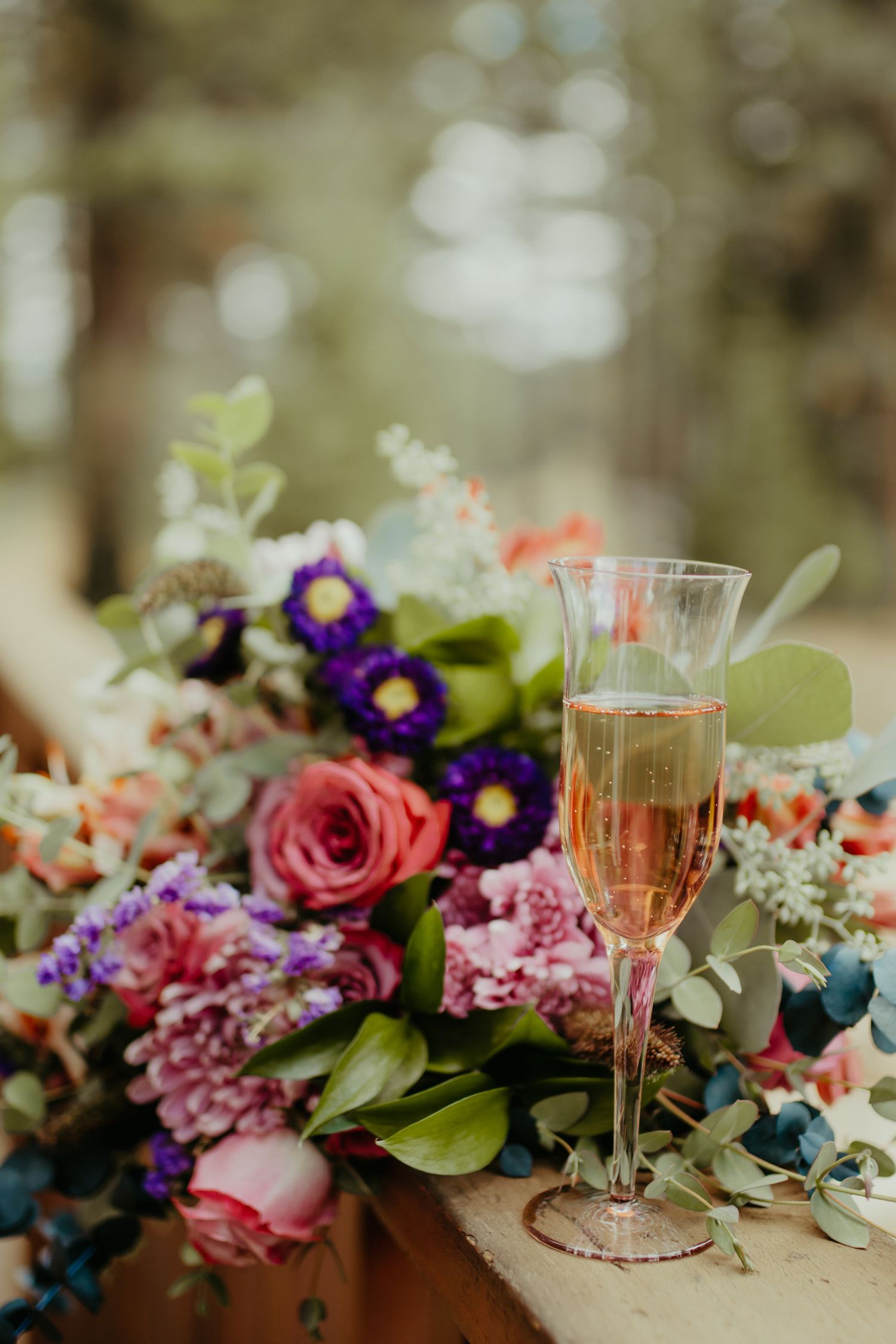 A champagne flute with rosé sits beside a colorful wedding bouquet of pink roses and purple flowers on a wooden railing.