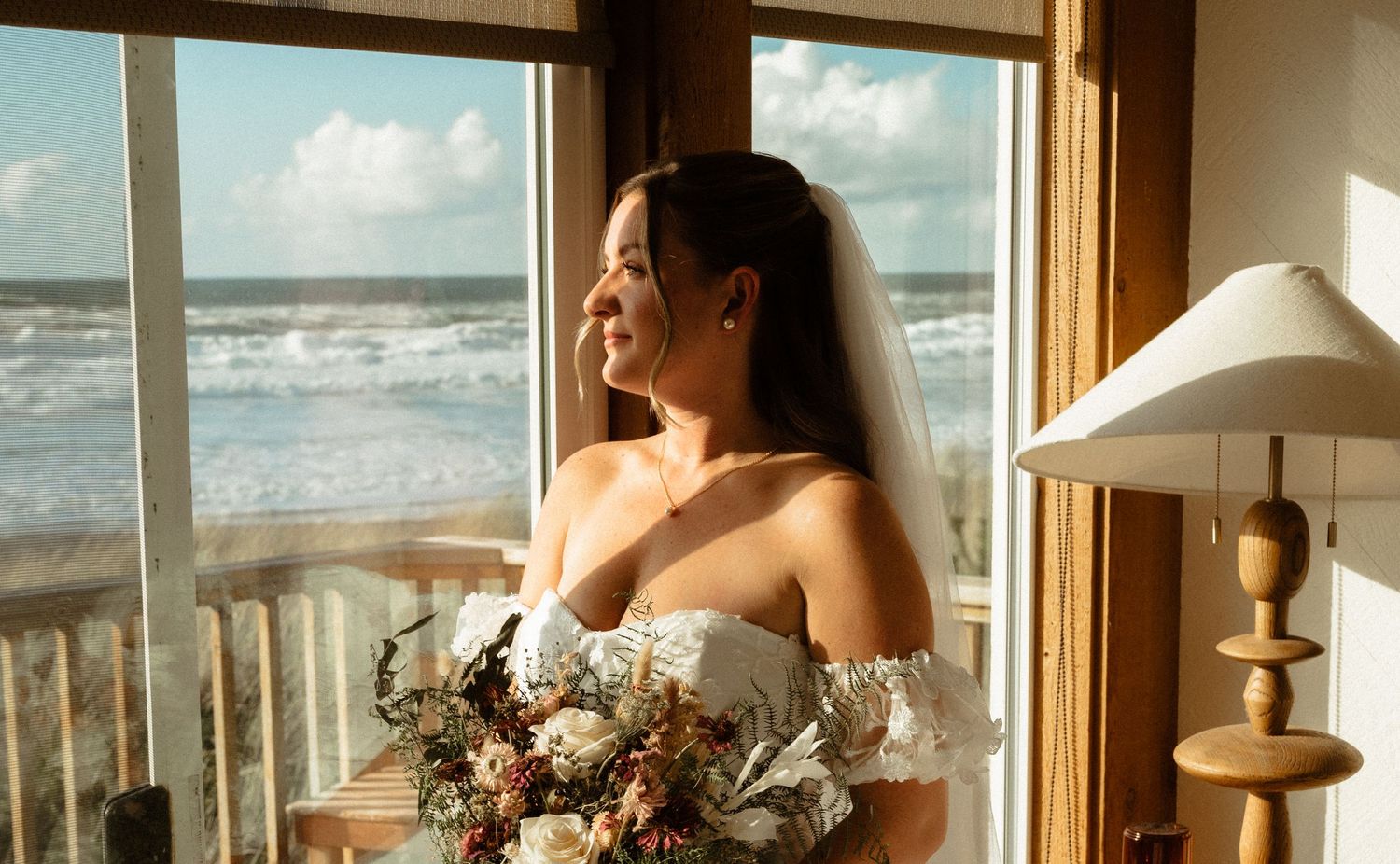 Bride in an off-the-shoulder white gown and veil holds a floral bouquet by a window with a sunlit ocean view.