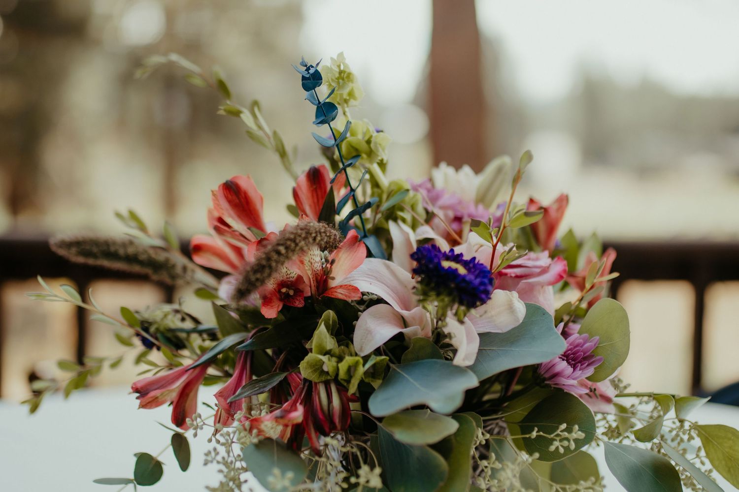 Colorful wedding bouquet with pink lilies, purple flowers, greenery, and feathers on a white surface.