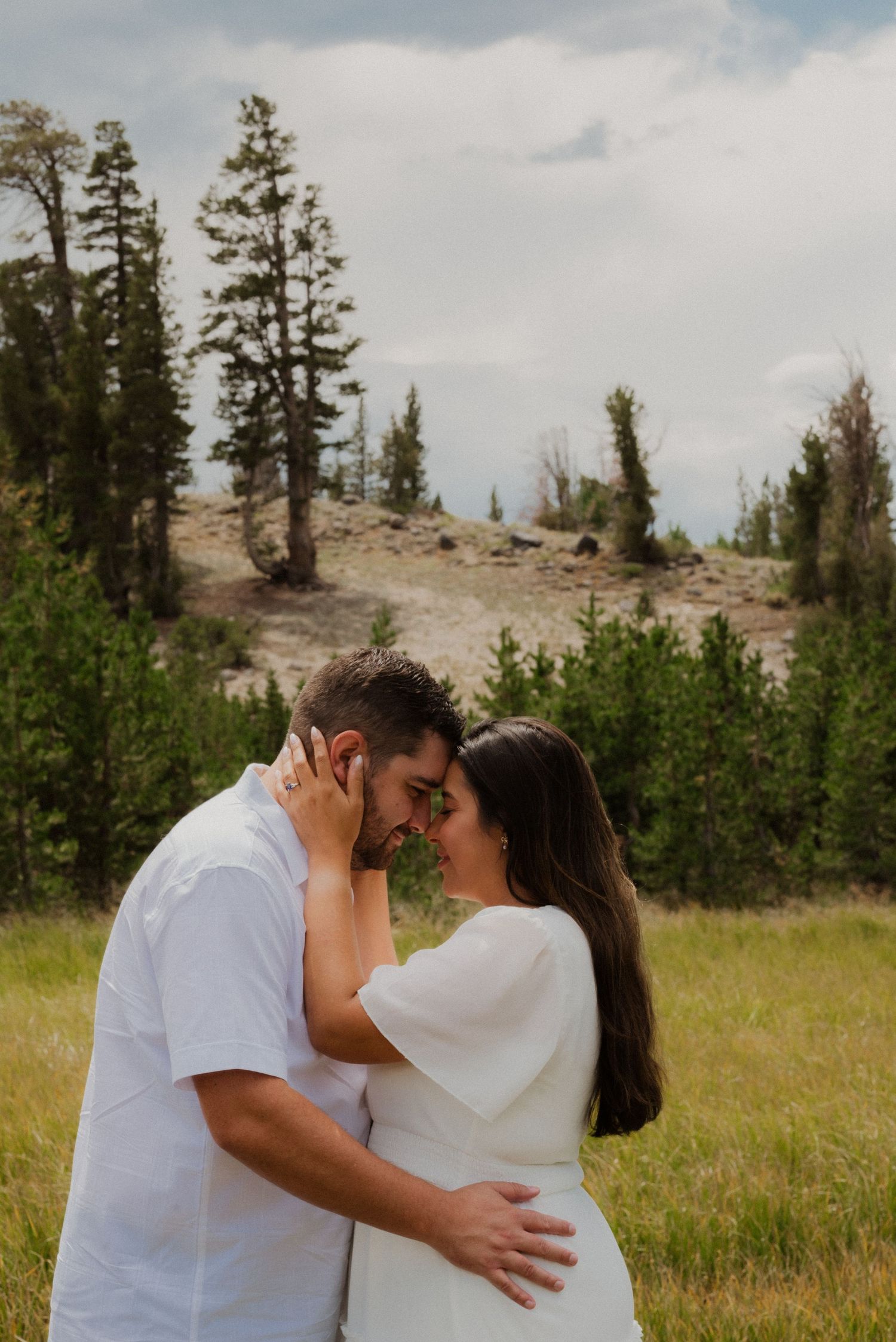 Couple in white outfits sharing a romantic moment in a lush green mountain meadow surrounded by pine trees.