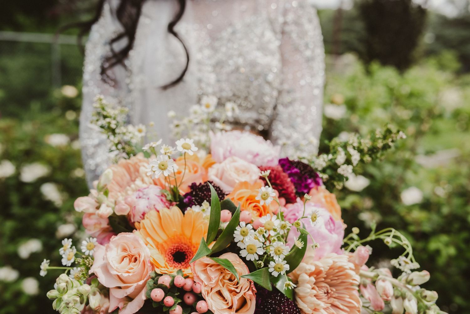 Bride in lace gown holding a vibrant wedding bouquet of peach, pink, and purple blooms with daisies in a garden.