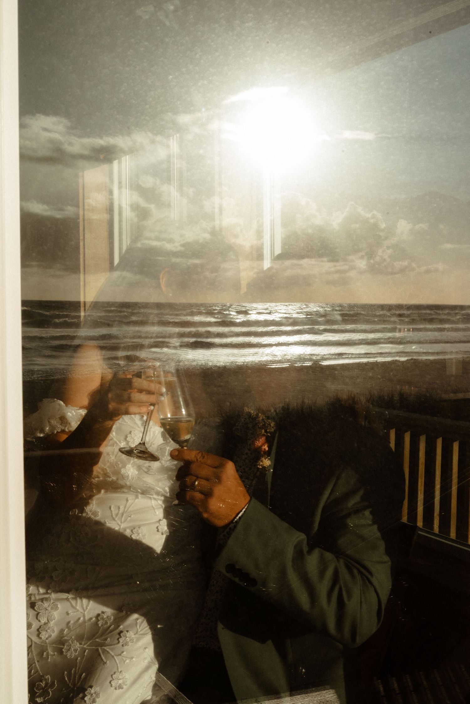 Newlyweds toasting champagne glasses by a sunlit ocean window, reflecting a beautiful beach sunset.