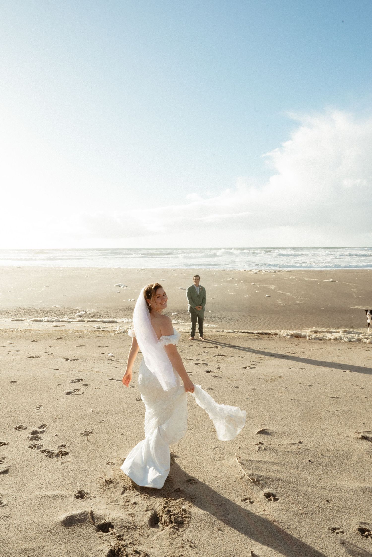 Bride in a flowing white gown and veil twirls on a sandy beach while the groom watches from a distance.