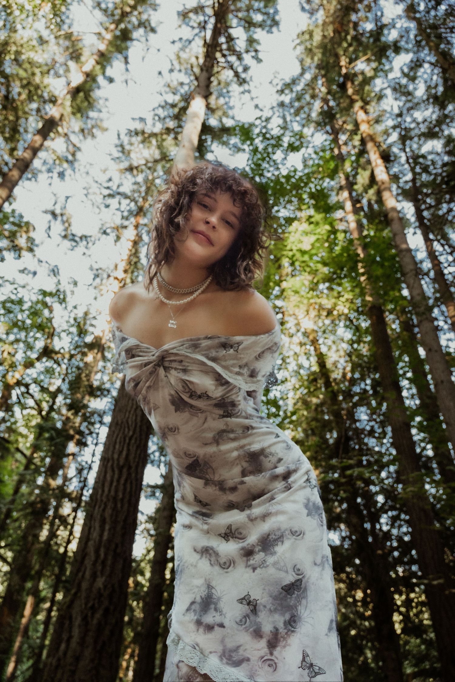 Woman in butterfly print off-shoulder dress posing in a sunlit forest, wearing layered pearl necklaces.
