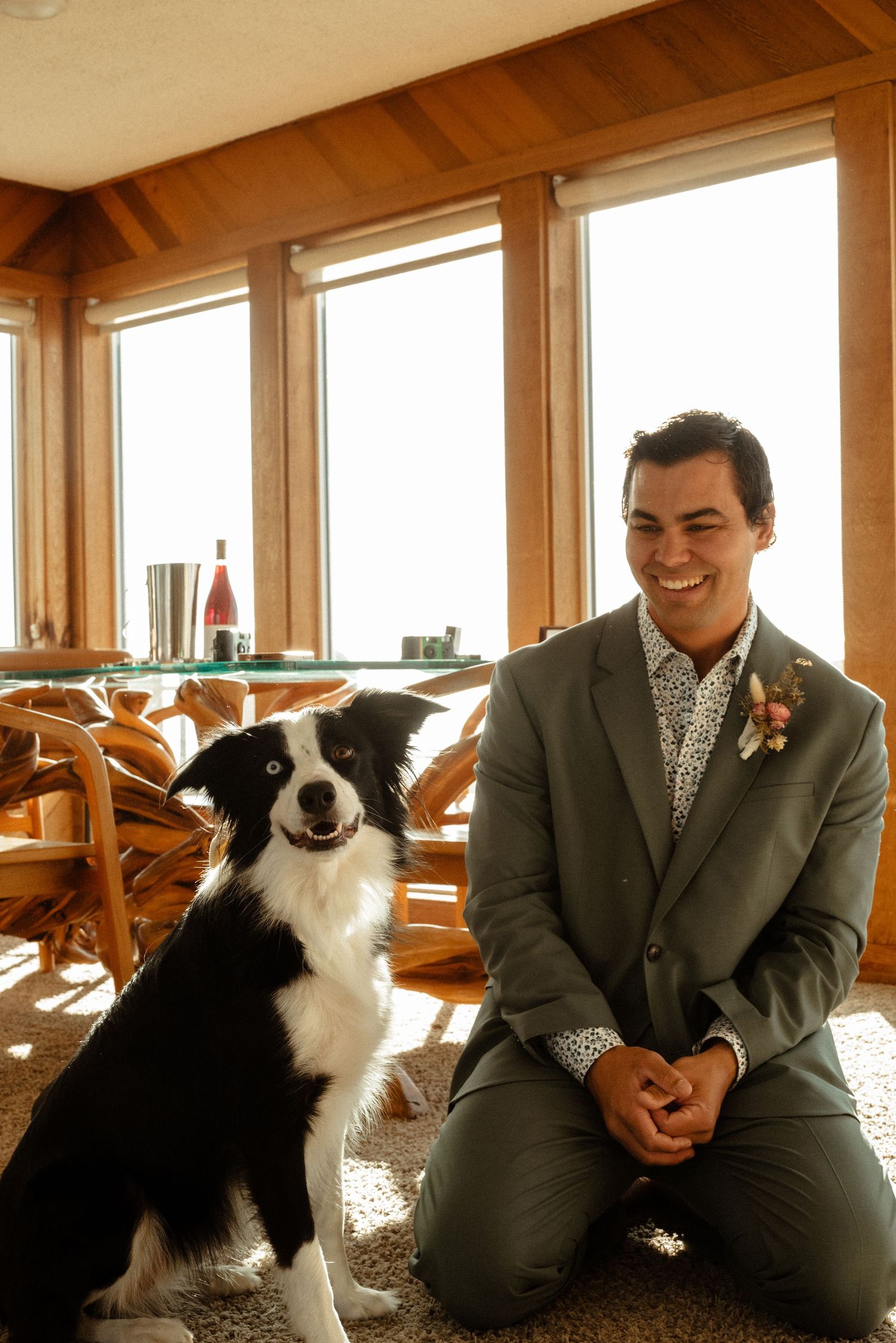 Smiling groom in a sage green suit with a floral boutonniere kneels beside his border collie in a sunlit beach house.