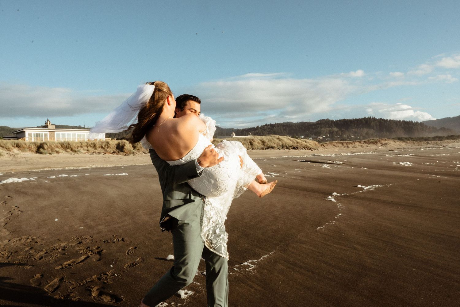 Groom in a gray suit carries his bride in a white wedding dress across a wide, wet sandy beach with a building and hills in the distance.