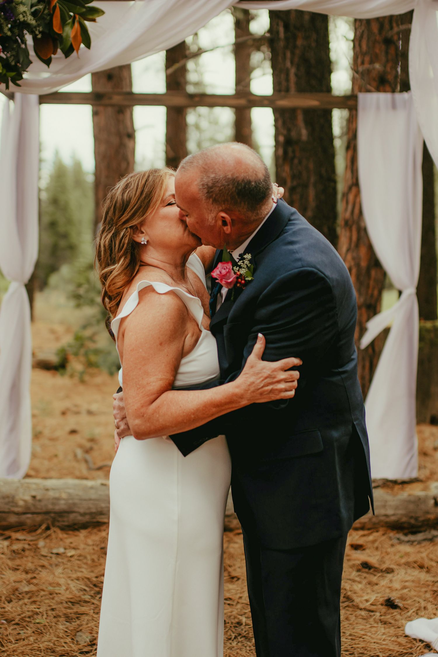 Bride and groom share their first kiss at an outdoor forest wedding ceremony with white draping and wooden arch.