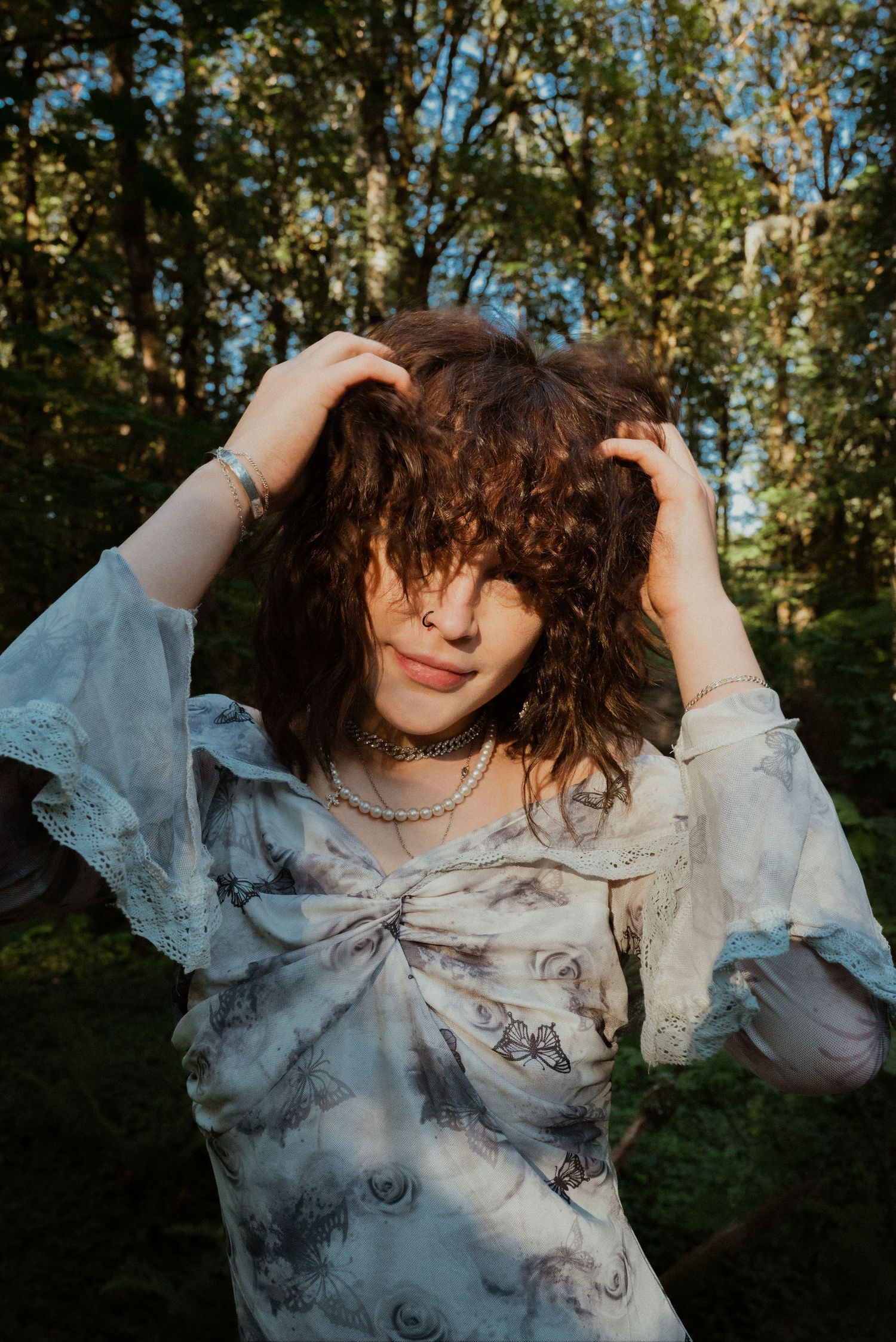 A young woman in a floral dress with lace sleeves holds her curly hair back in a sunlit forest setting.