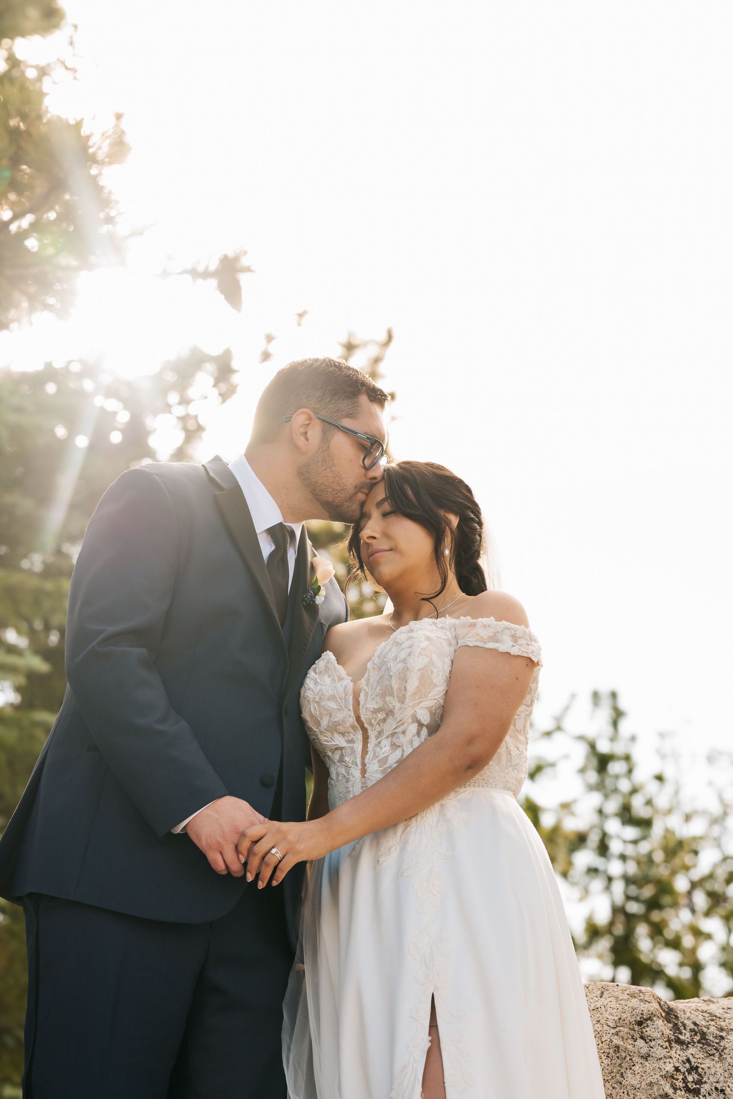 Bride and groom share a romantic moment outdoors, bathed in golden sunlight, with pine trees in the background.