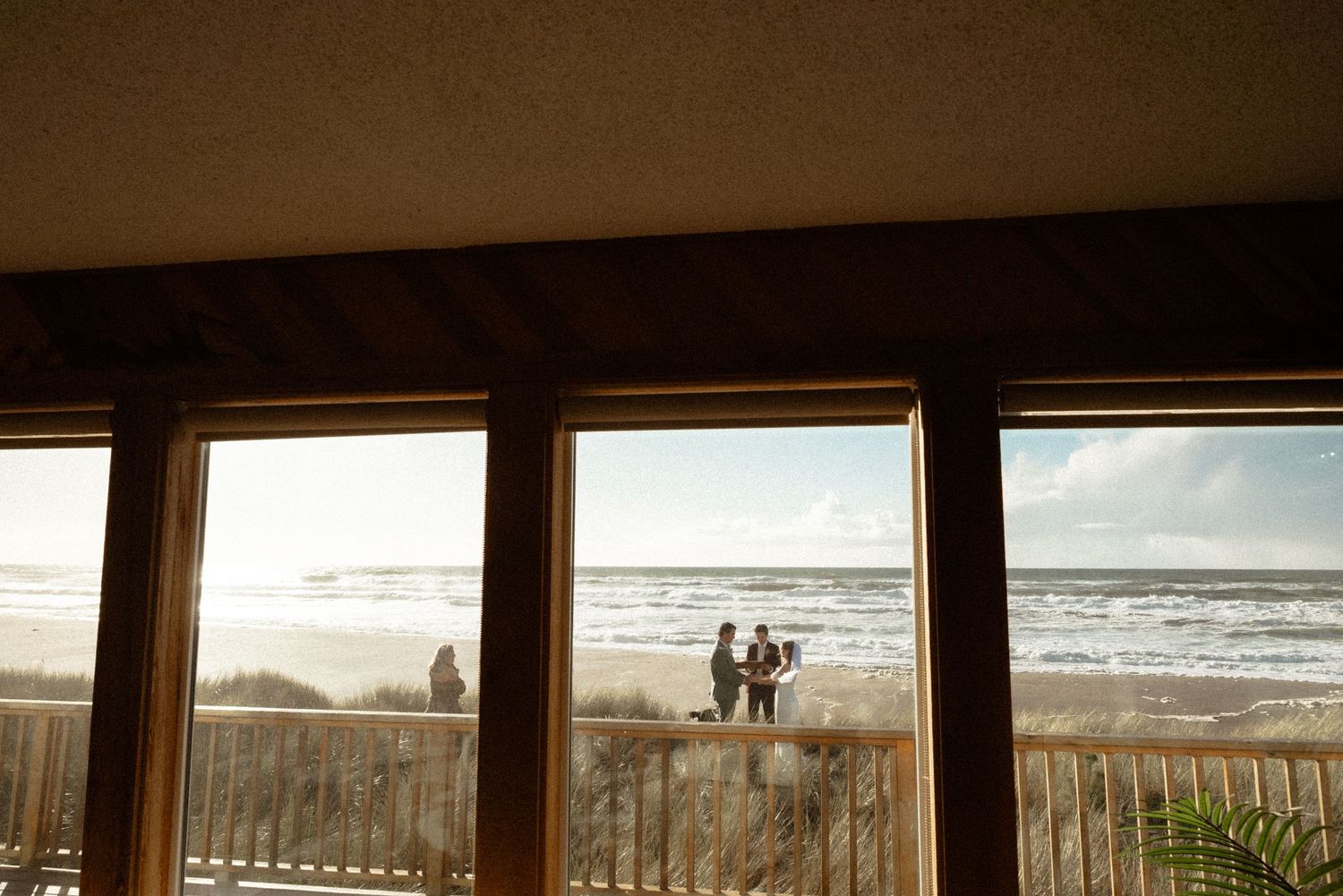 View through large windows of a small beach wedding ceremony on the sand, with ocean waves and a wooden deck railing.