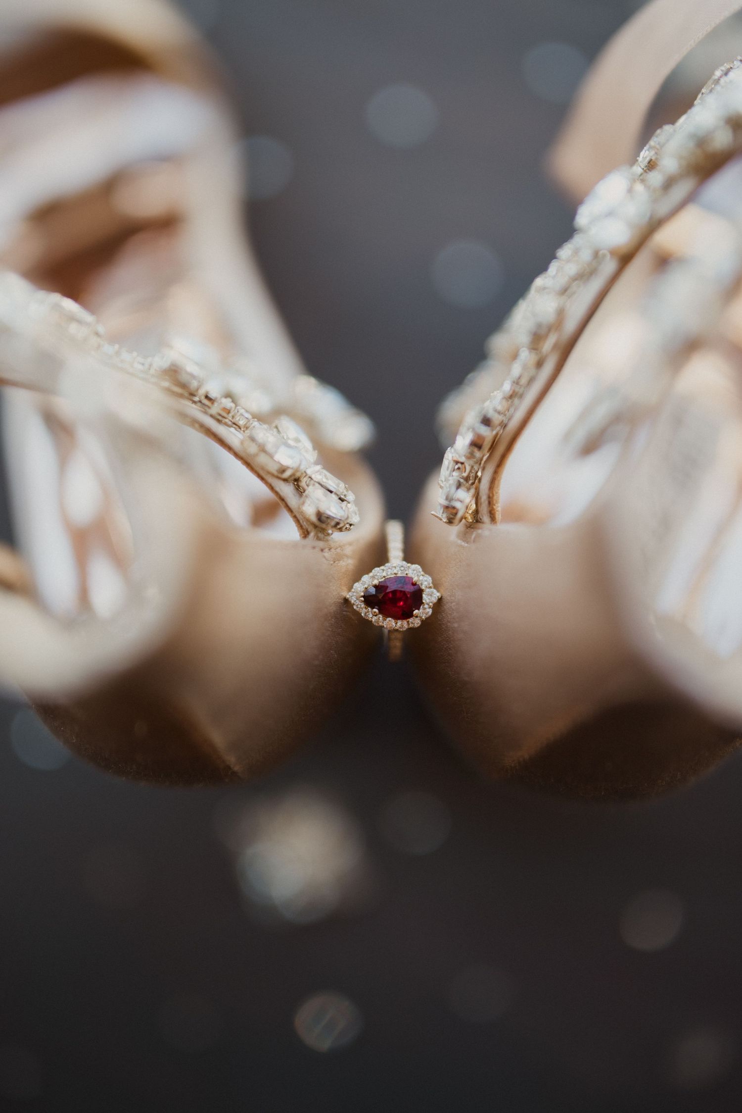 A ruby and diamond halo engagement ring resting between two ivory satin bridal heels with pearl embellishments.