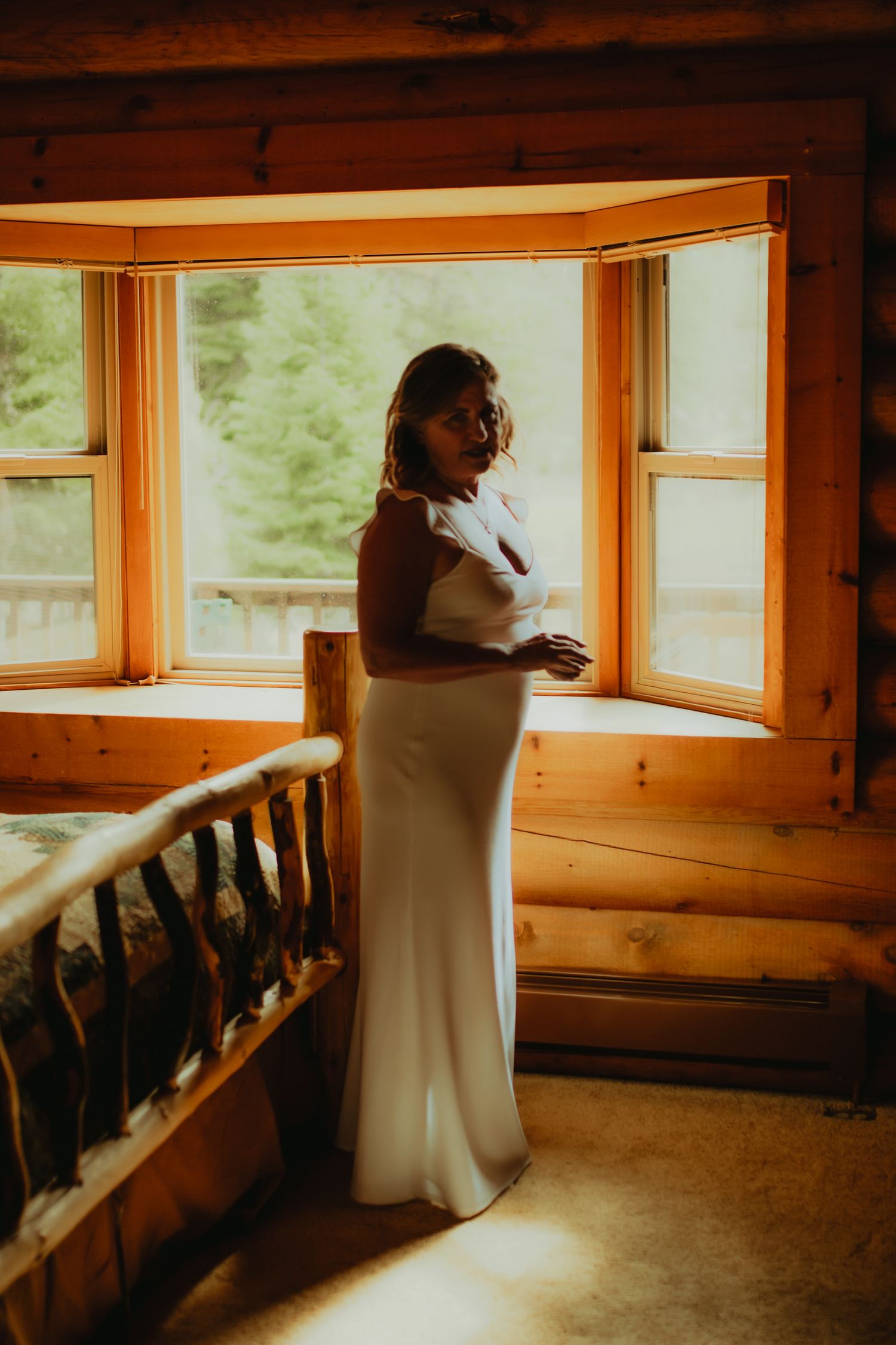 Bride in white gown standing by bay windows in rustic log cabin, bathed in warm golden light.