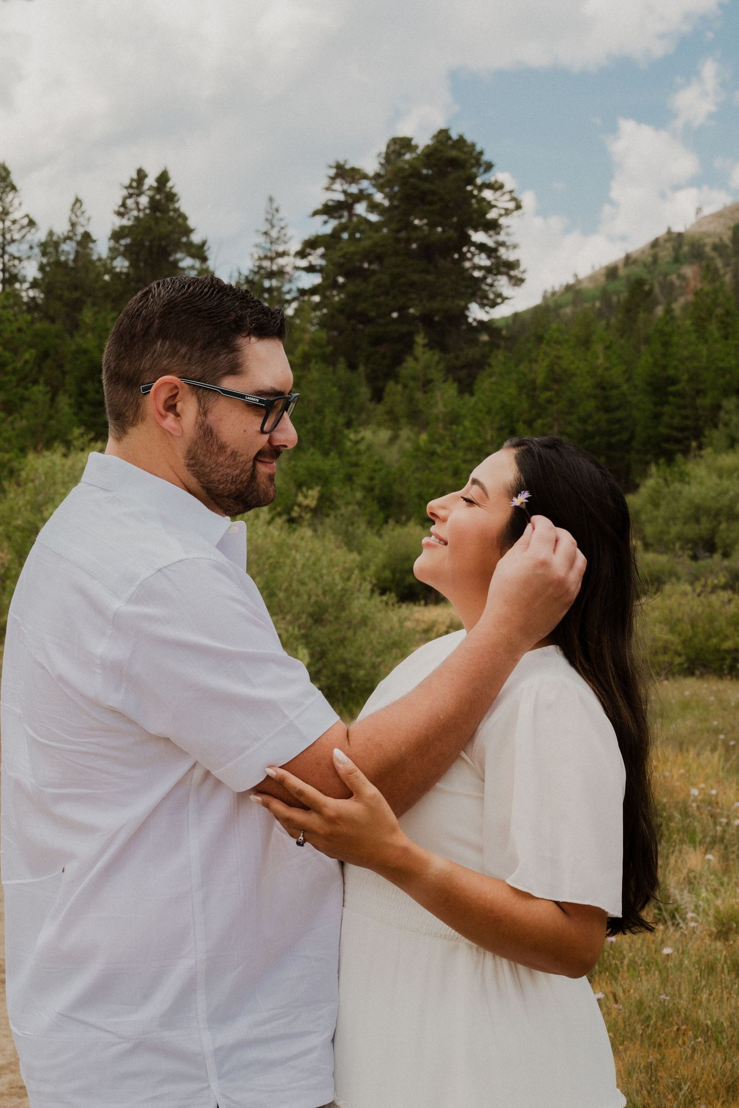 Couple in white outfits sharing a tender moment in a mountain meadow with pine trees and cloudy sky.
