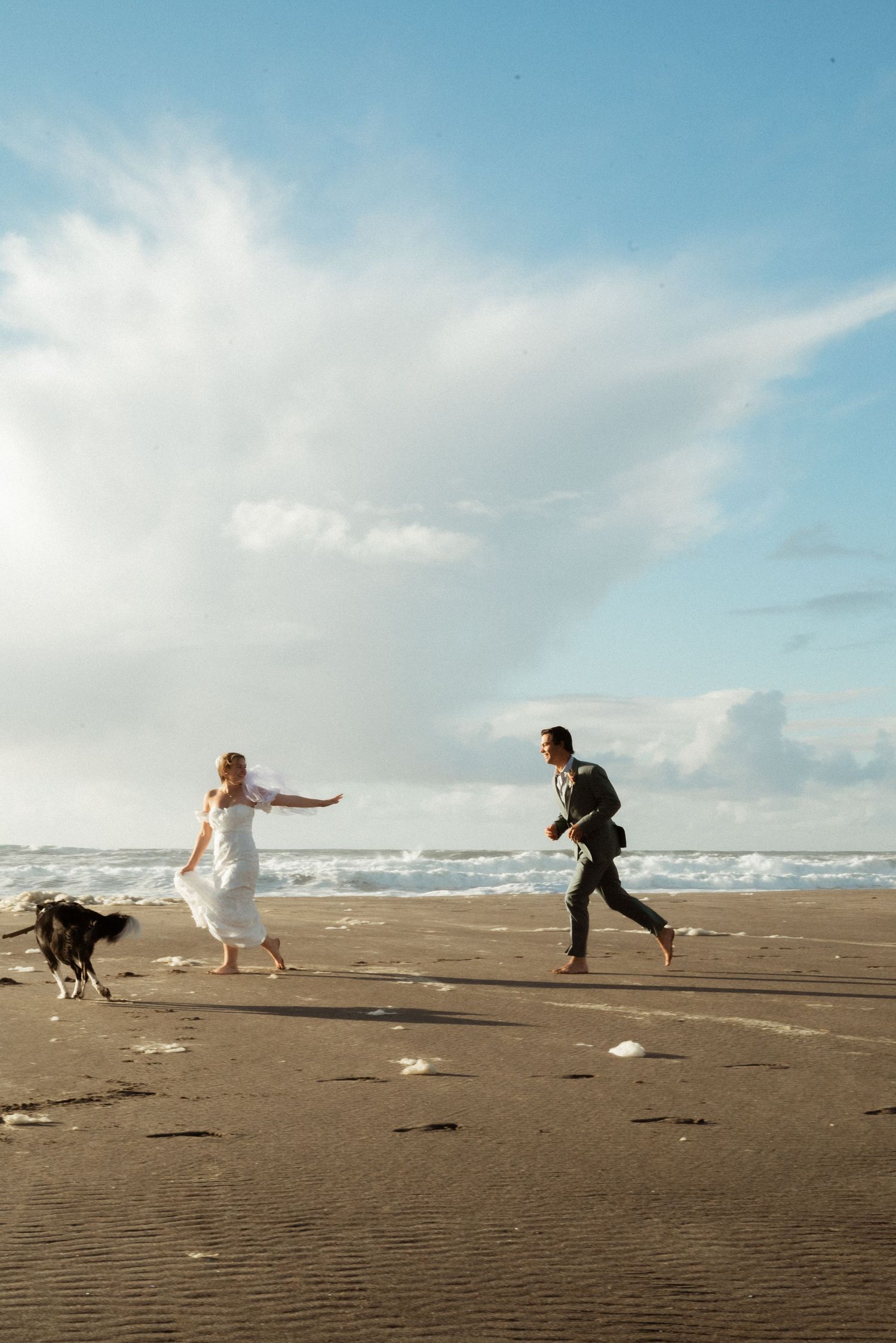 Bride in a white dress and groom in a dark suit run barefoot along a sandy beach with a black and white dog under a bright blue sky.