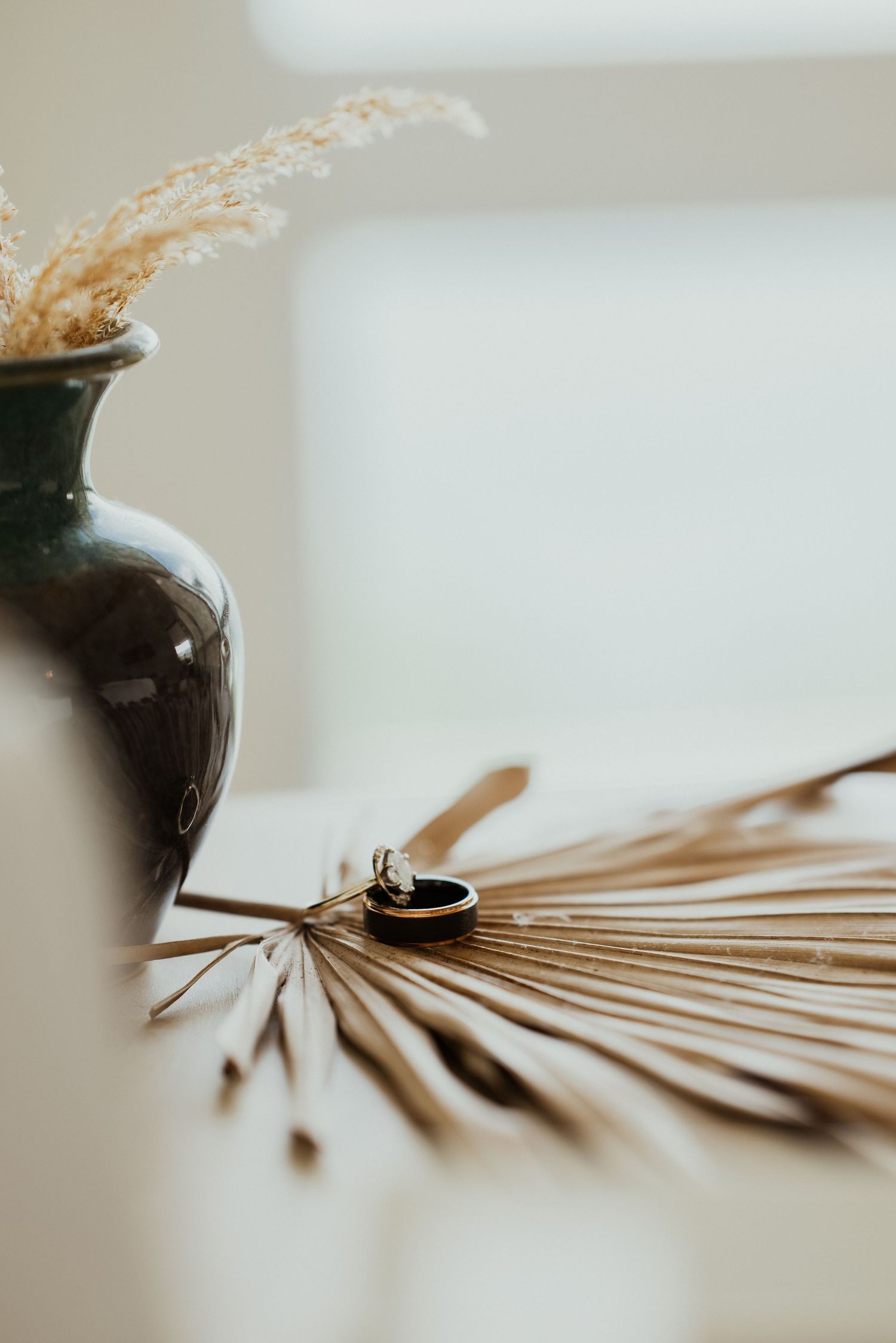 Wedding rings rest on dried palm leaf beside a green ceramic vase with dried florals.
