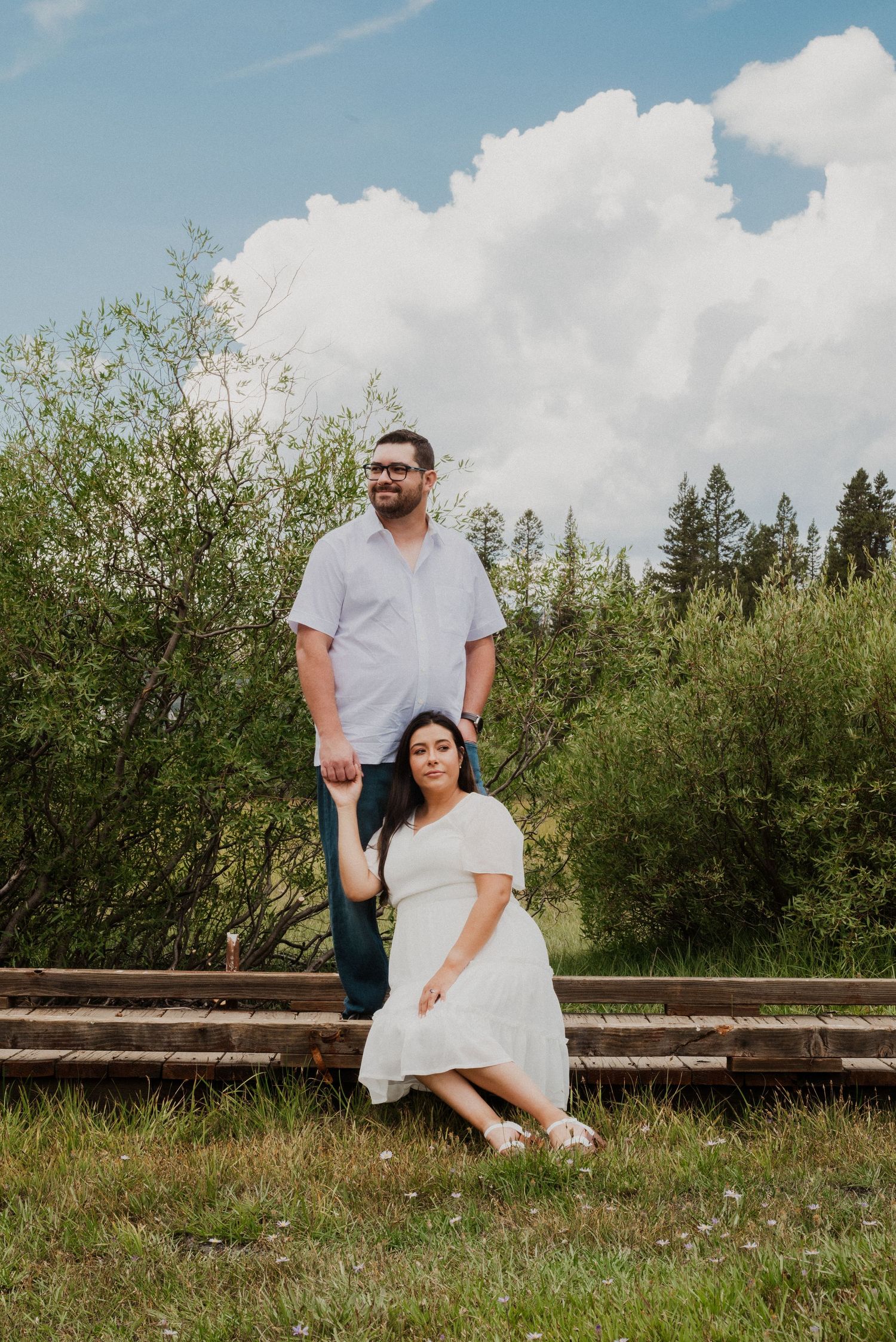 Couple posing outdoors on railroad tracks, woman in white dress seated, man standing behind, lush green trees and clouds.