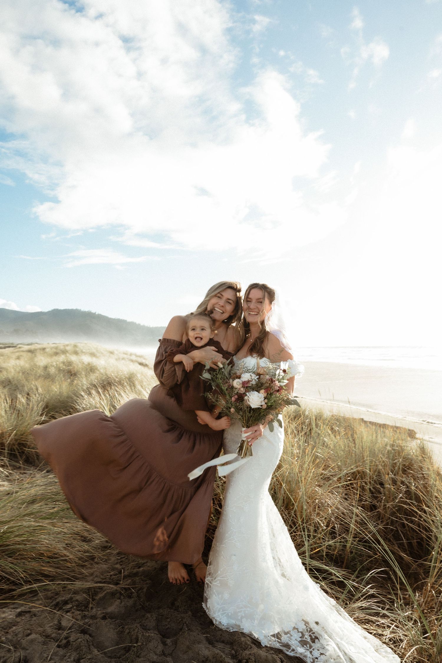 Bride in a lace gown holding a bouquet poses with a woman and toddler among dune grass on a sunny beach.
