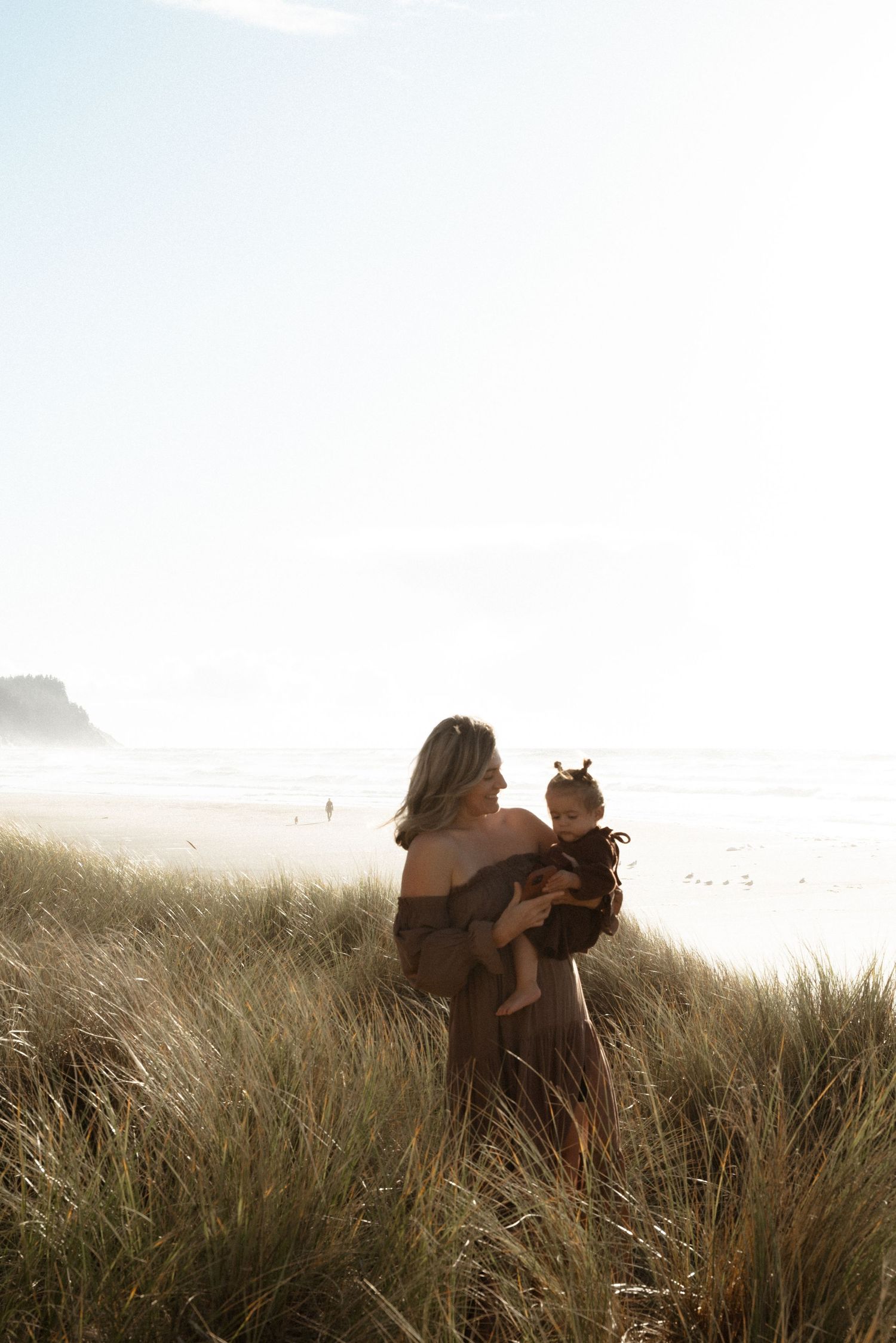 A woman in an off-the-shoulder dress holds a toddler while standing in tall beach grass with a hazy ocean and sandy shoreline behind them.