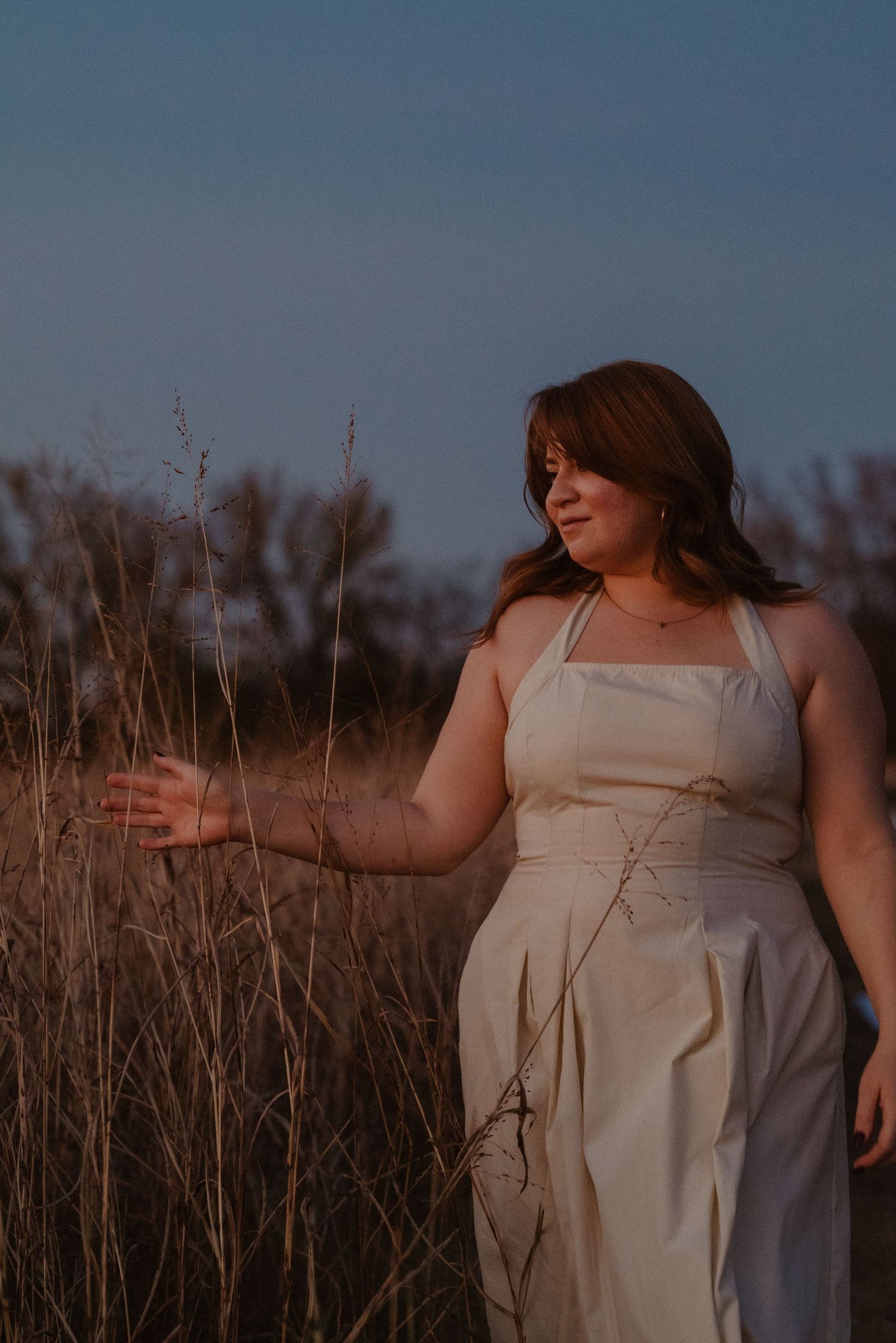 Woman in white halter dress touching tall grass at golden hour in a field.