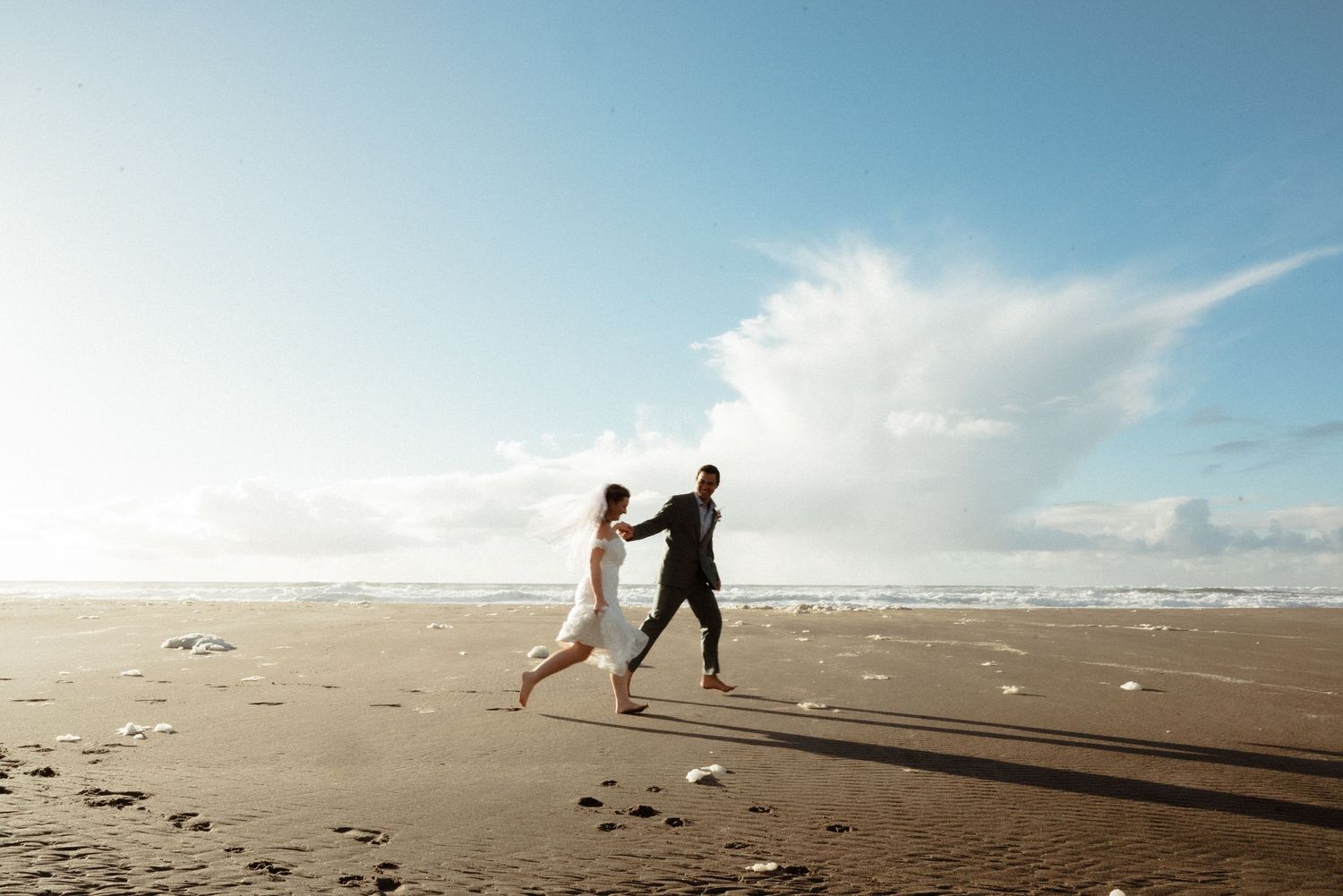 Bride and groom running joyfully on a sandy beach with ocean waves and blue sky backdrop.