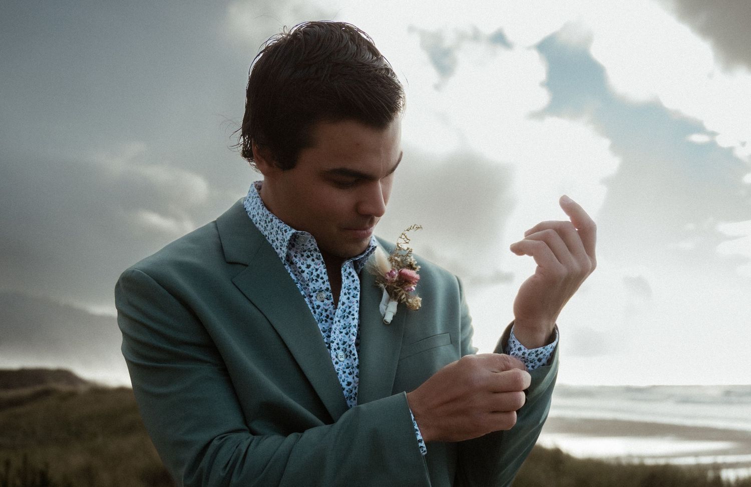 Groom in green suit adjusting cufflinks outdoors with boutonniere, dramatic cloudy sky and beach backdrop.