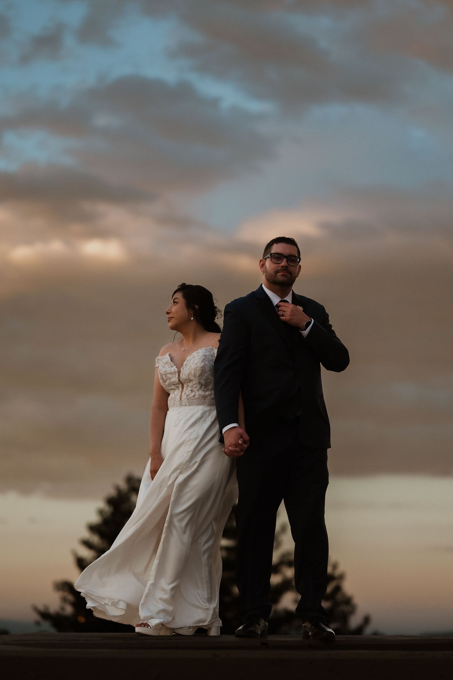 Bride and groom hold hands at sunset, dramatic cloudy sky backdrop, wedding portrait outdoors.