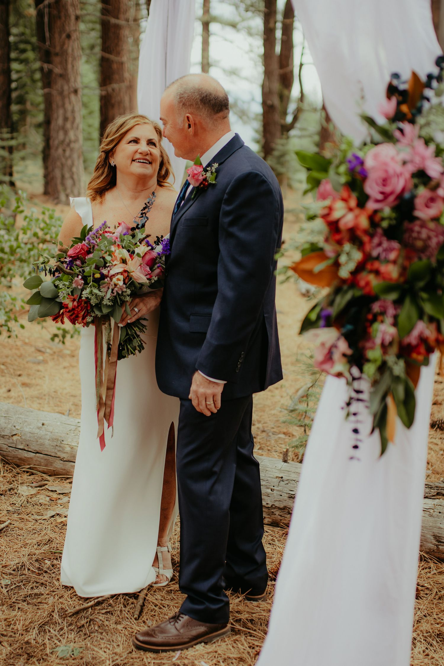 Bride and groom share a tender moment at outdoor forest wedding ceremony with colorful floral bouquet.