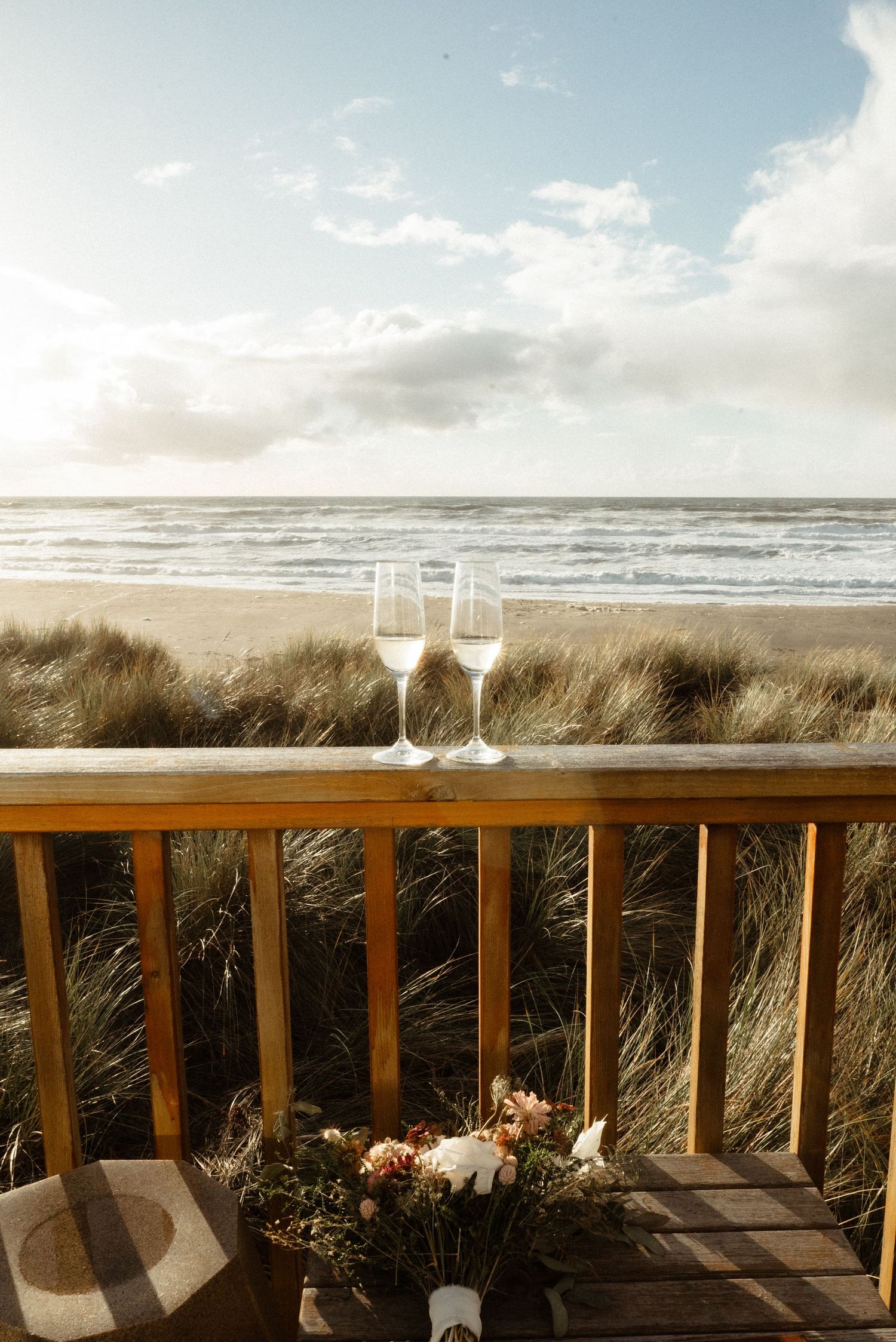 Two champagne glasses and a small floral bouquet sit on a wooden deck railing overlooking beach grass and ocean waves.