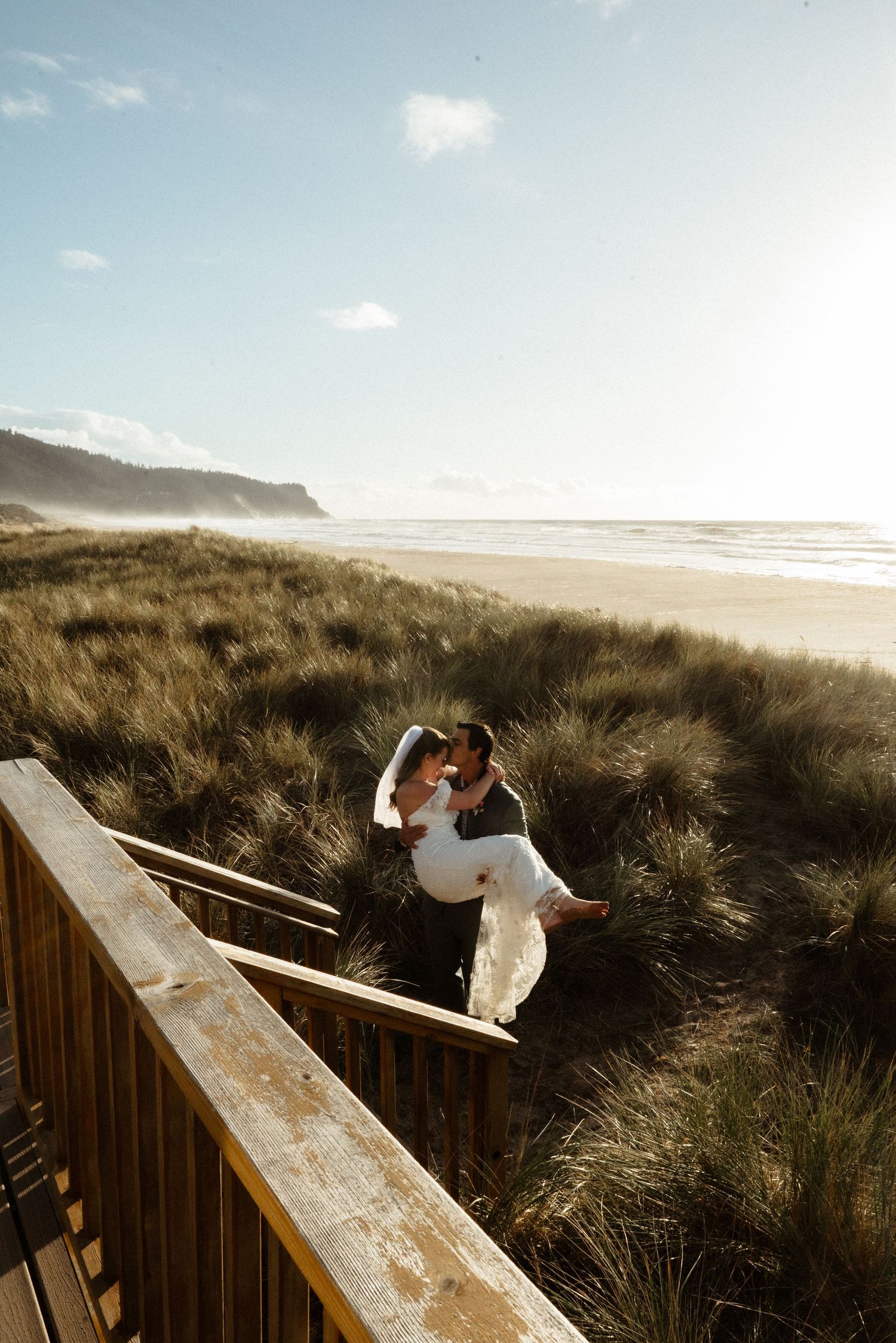 A groom carries his bride in a white wedding dress down wooden stairs through beach grass toward a sunlit ocean shore.