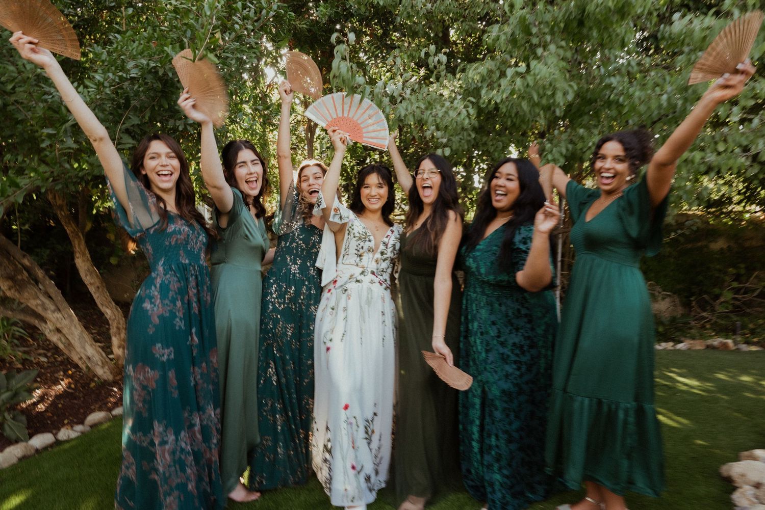 Bride in white floral gown celebrates with six bridesmaids in green dresses, holding fans outdoors.