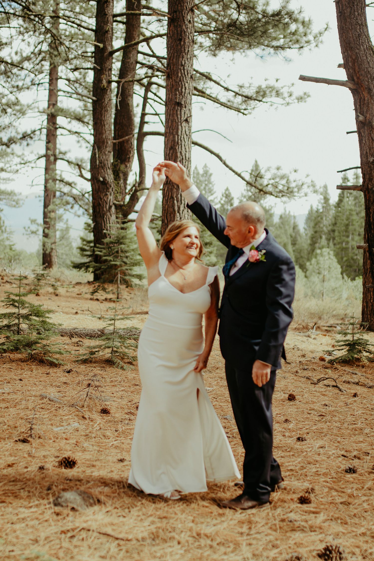 Bride and groom dancing joyfully outdoors in a pine forest, wearing wedding attire with pine cones on the ground.