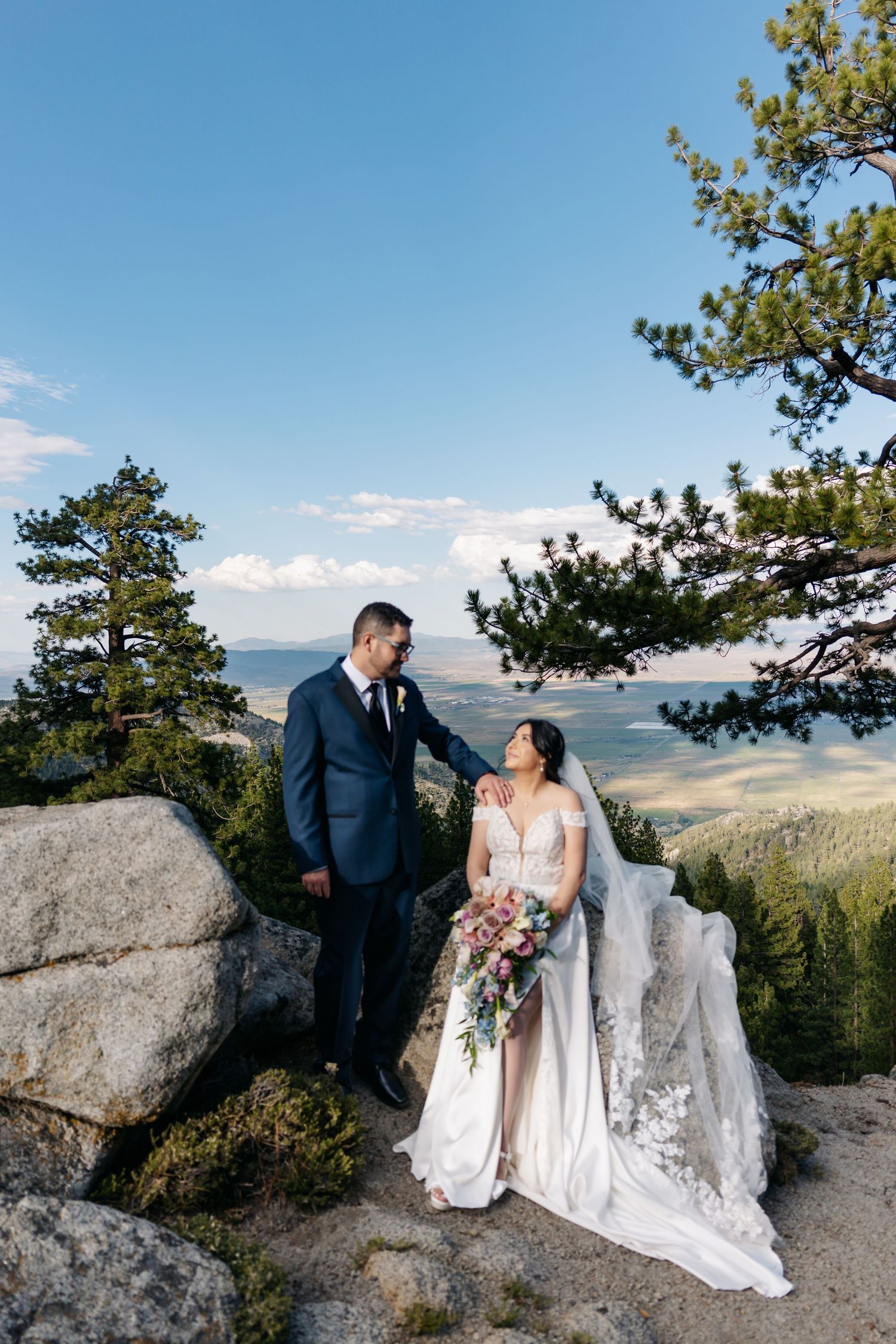 Bride and groom pose on rocky mountain overlook with panoramic valley views, pine trees, and blue sky backdrop.