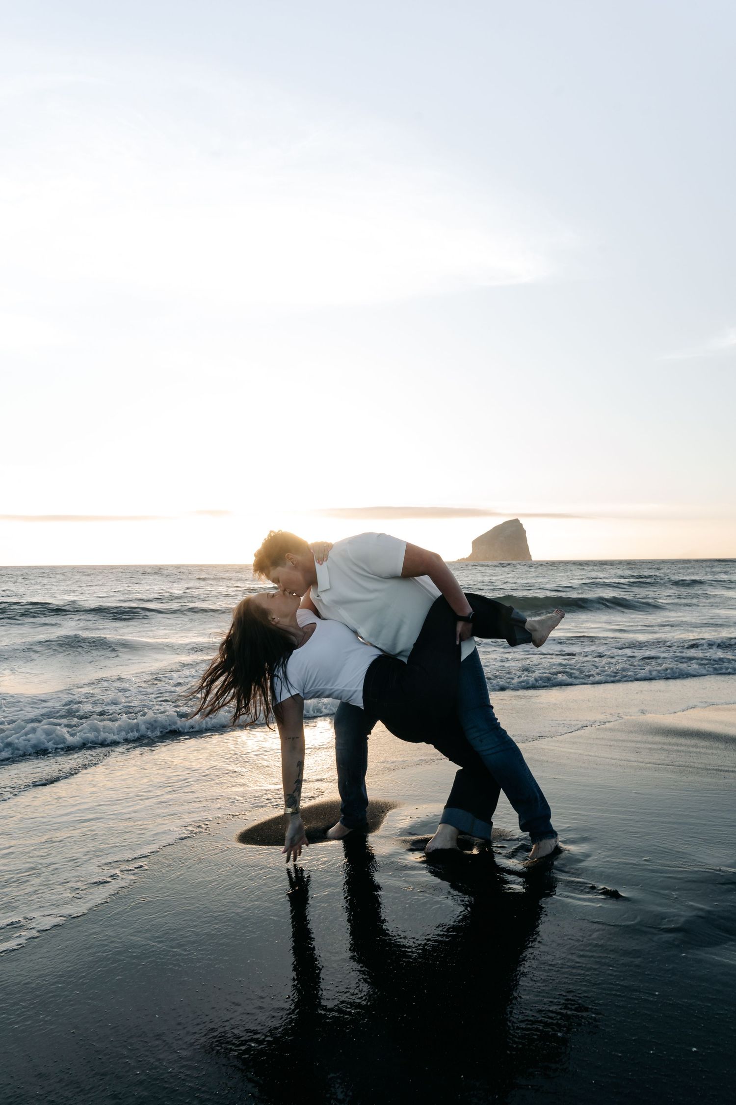 Couple sharing a romantic dip kiss on a wet black sand beach at sunset with ocean waves and rock formation behind.