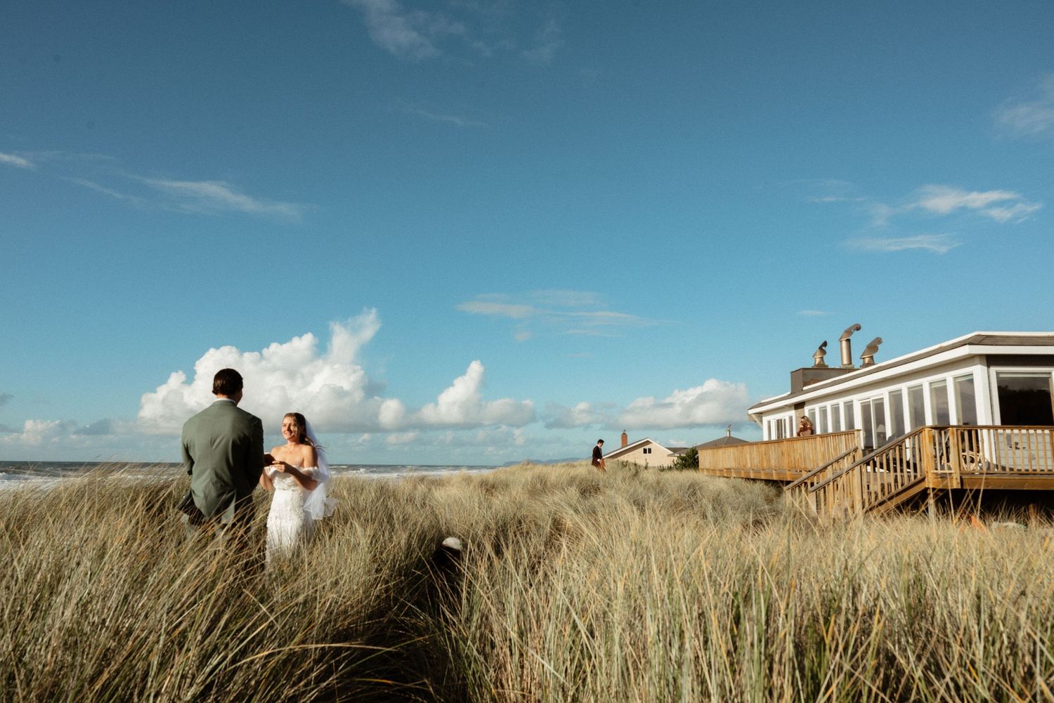 Bride and groom stand together in tall beach grass beneath a blue sky near a seaside building on their wedding day.