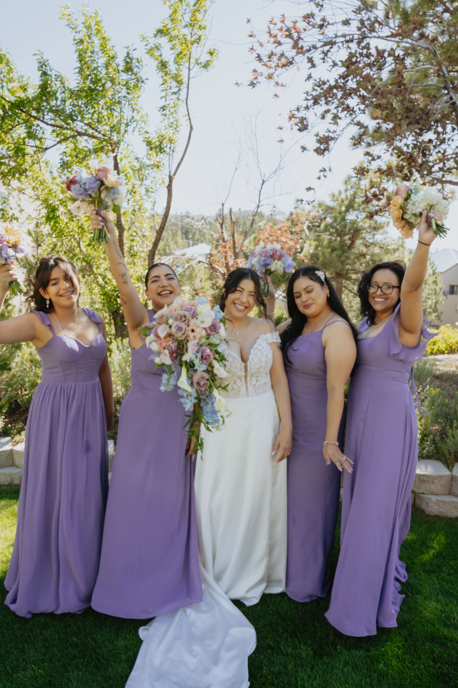 Bride in white gown with four bridesmaids in purple dresses raising bouquets outdoors.