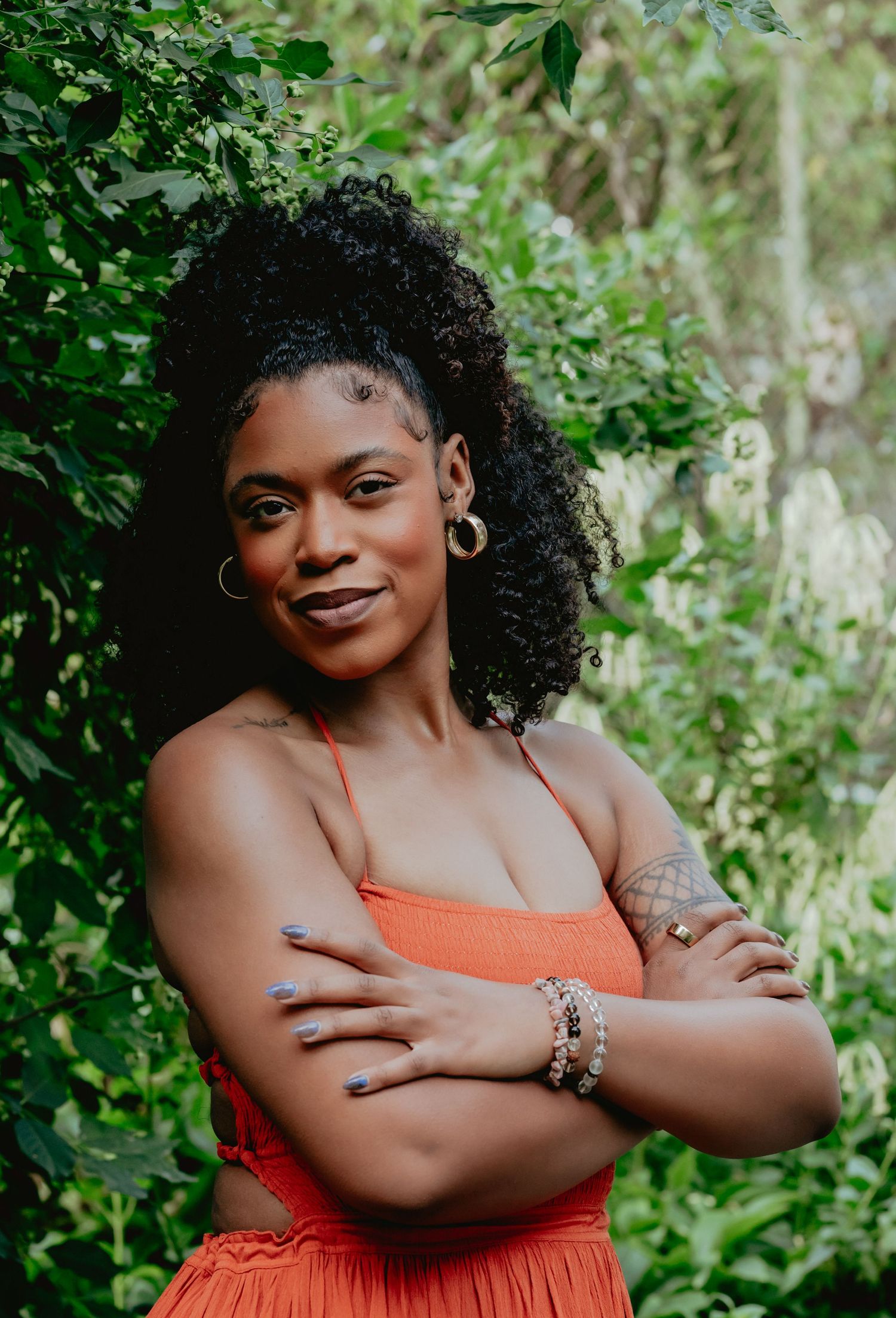 A confident Black woman in an orange outfit poses with arms crossed among lush green foliage.