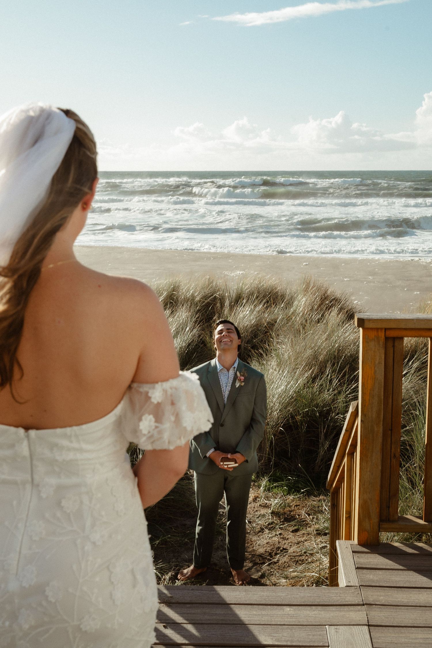 Bride in a white lace gown faces a groom in a gray suit who looks up emotionally on a wooden beach staircase overlooking the ocean.