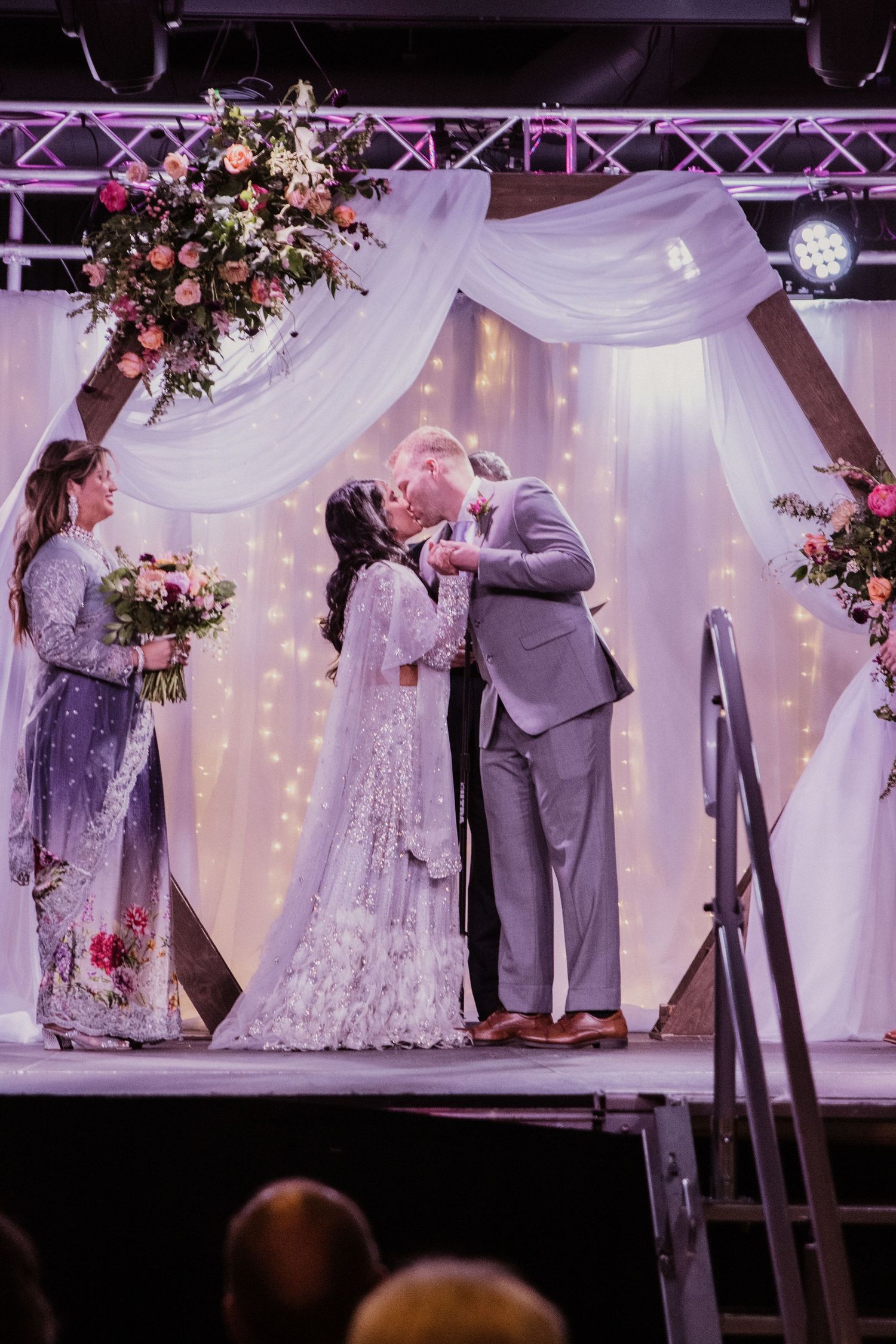 Bride and groom share first kiss at elegant wedding altar with white draping, fairy lights, and pink floral arrangements.