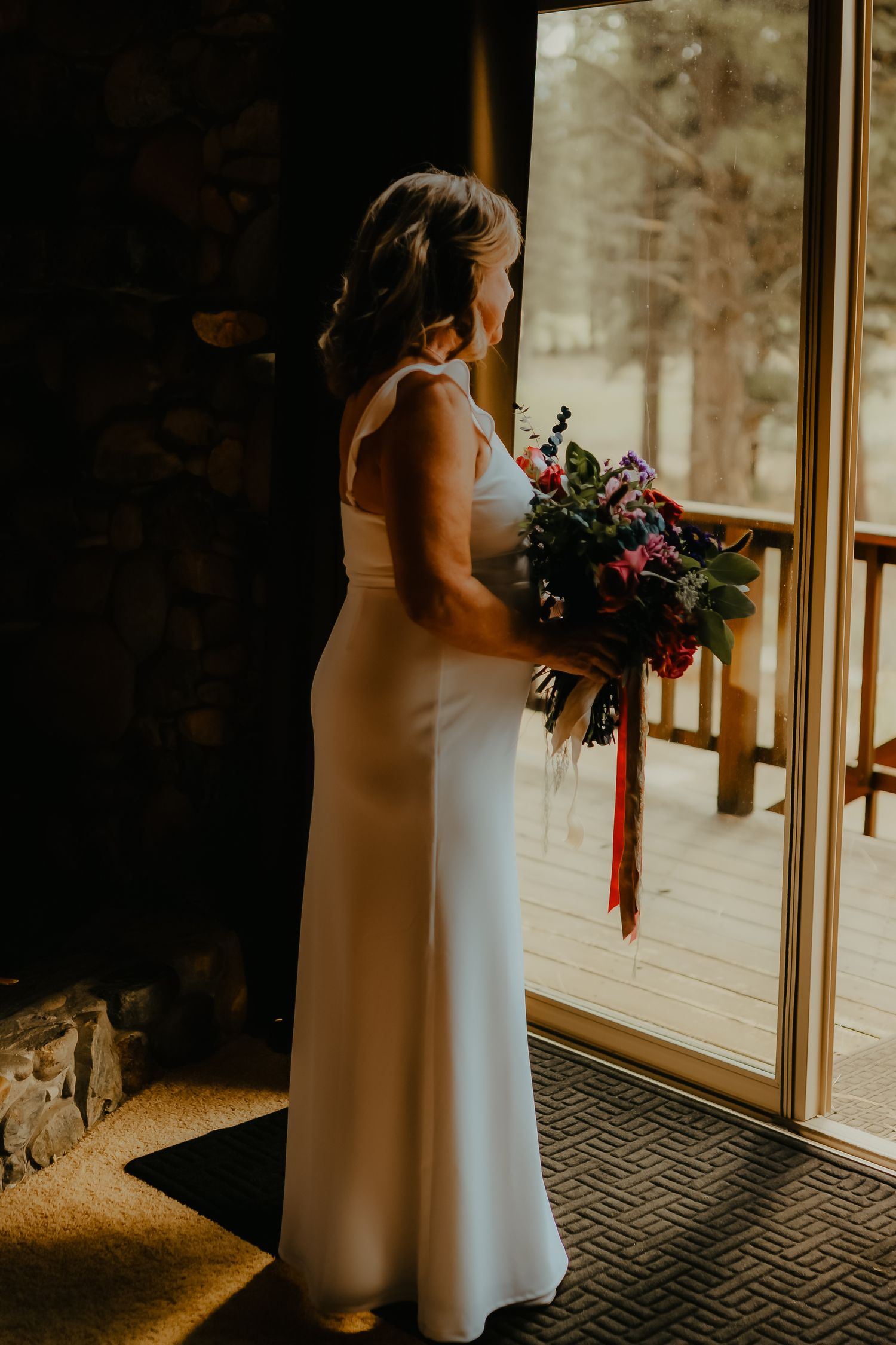Bride in white gown holding colorful bouquet stands by glass door in rustic cabin wedding venue.