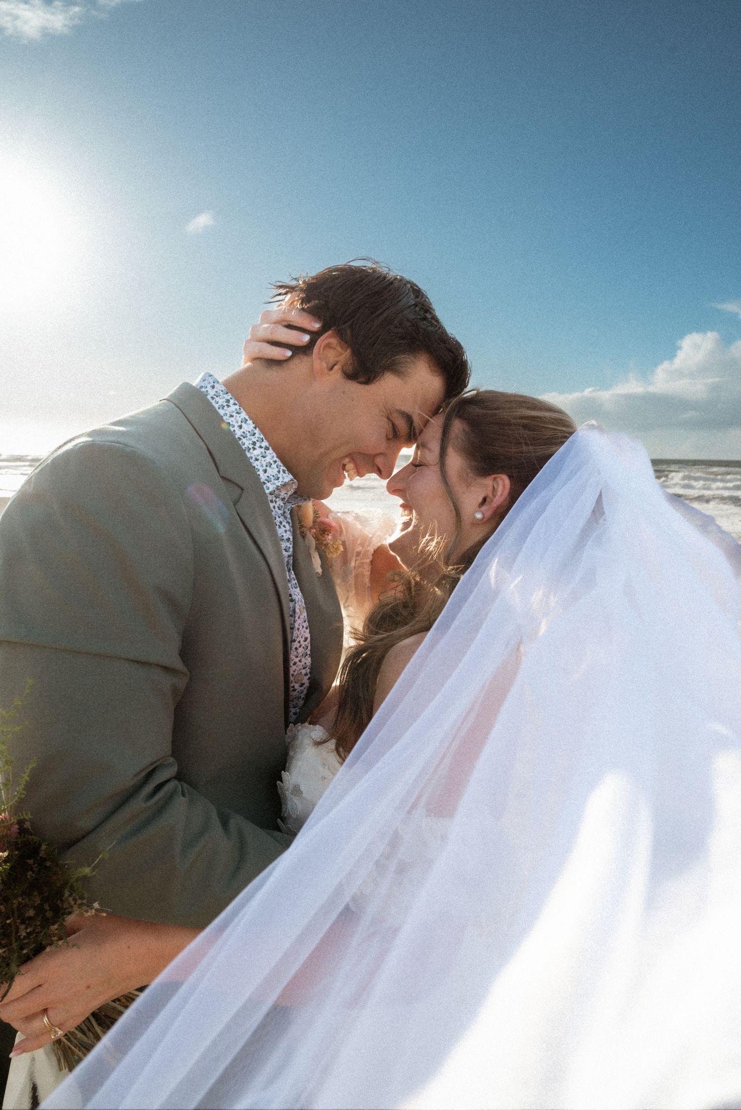 Bride and groom touch foreheads in a tender moment on the beach as her sheer veil drifts in the ocean breeze.