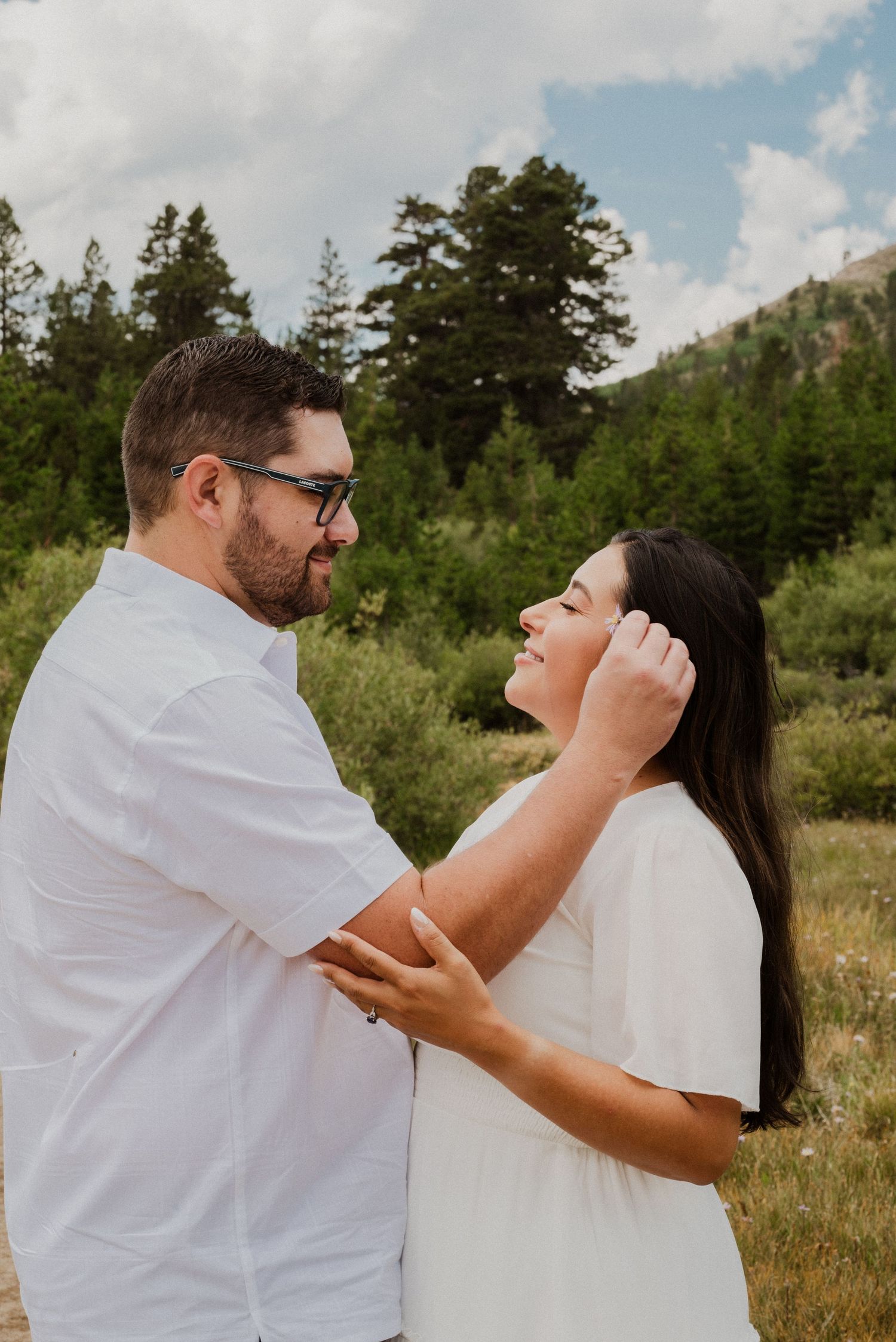 Couple in matching white outfits share a tender moment in a mountain meadow surrounded by pine trees.