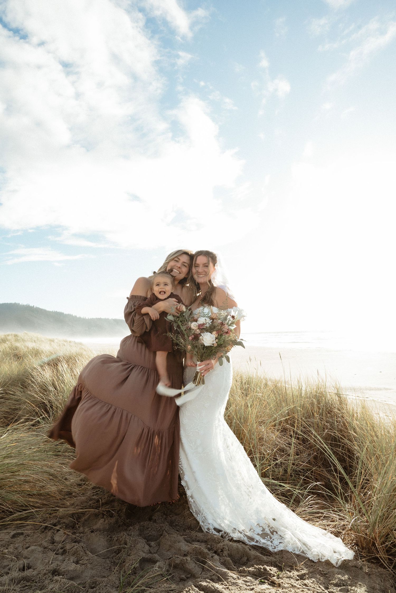Bride in lace gown holds bouquet with woman in brown dress and child on sandy beach dunes.