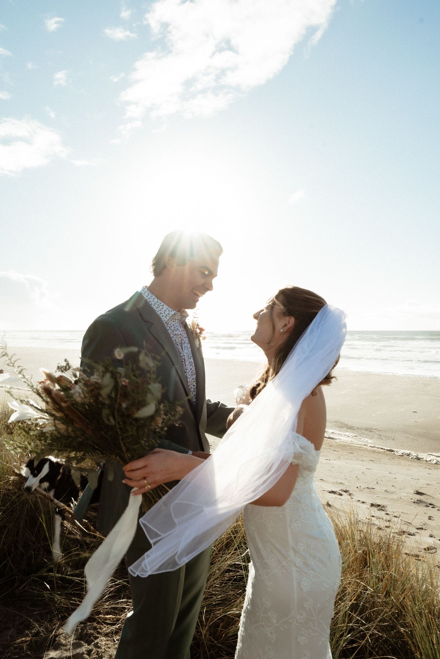 Bride and groom share a joyful moment on a sunlit beach, her veil blowing in the breeze as he holds a rustic bridal bouquet.