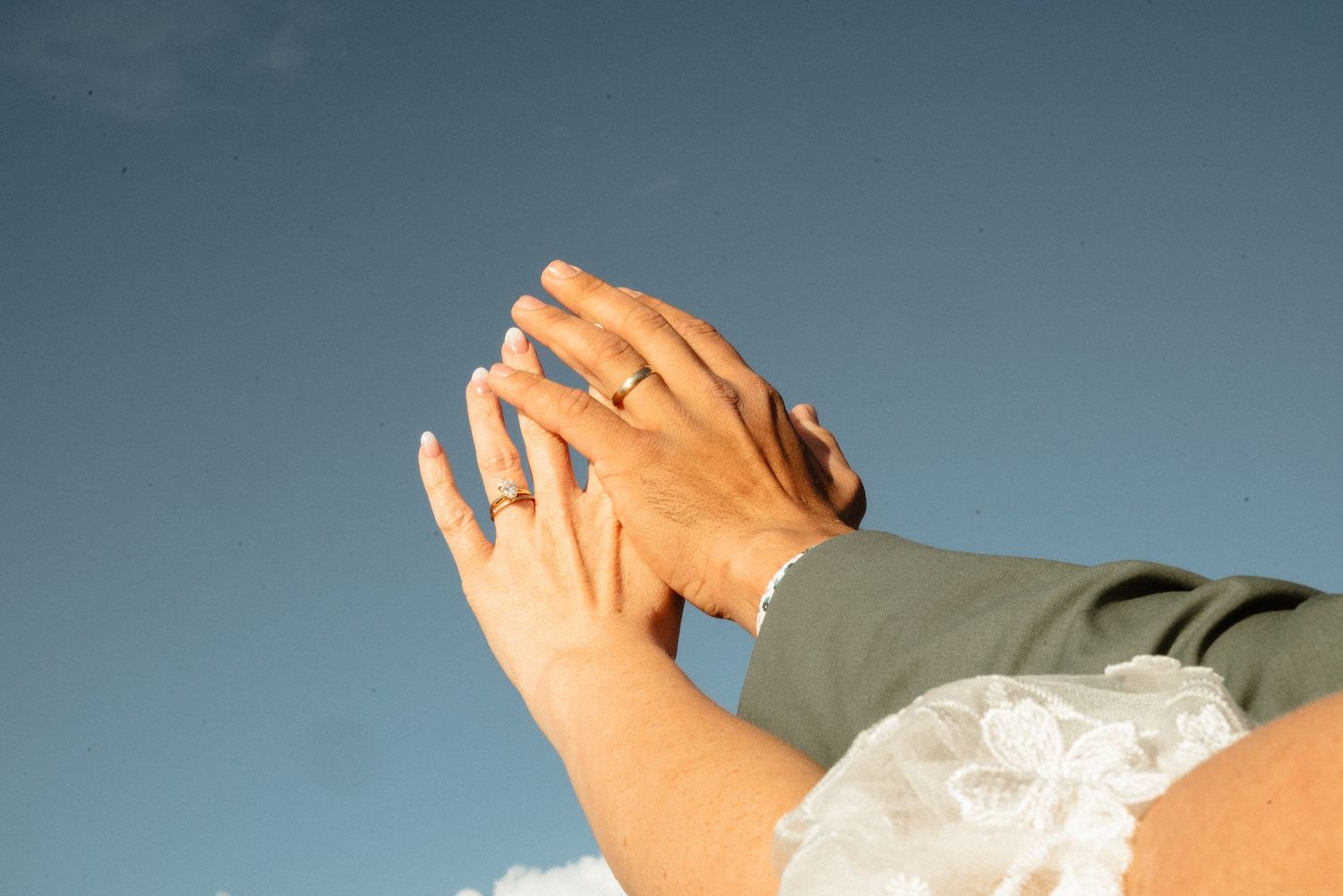 Couple's hands with wedding rings reach together against a clear blue sky, the bride's lace sleeve visible nearby.